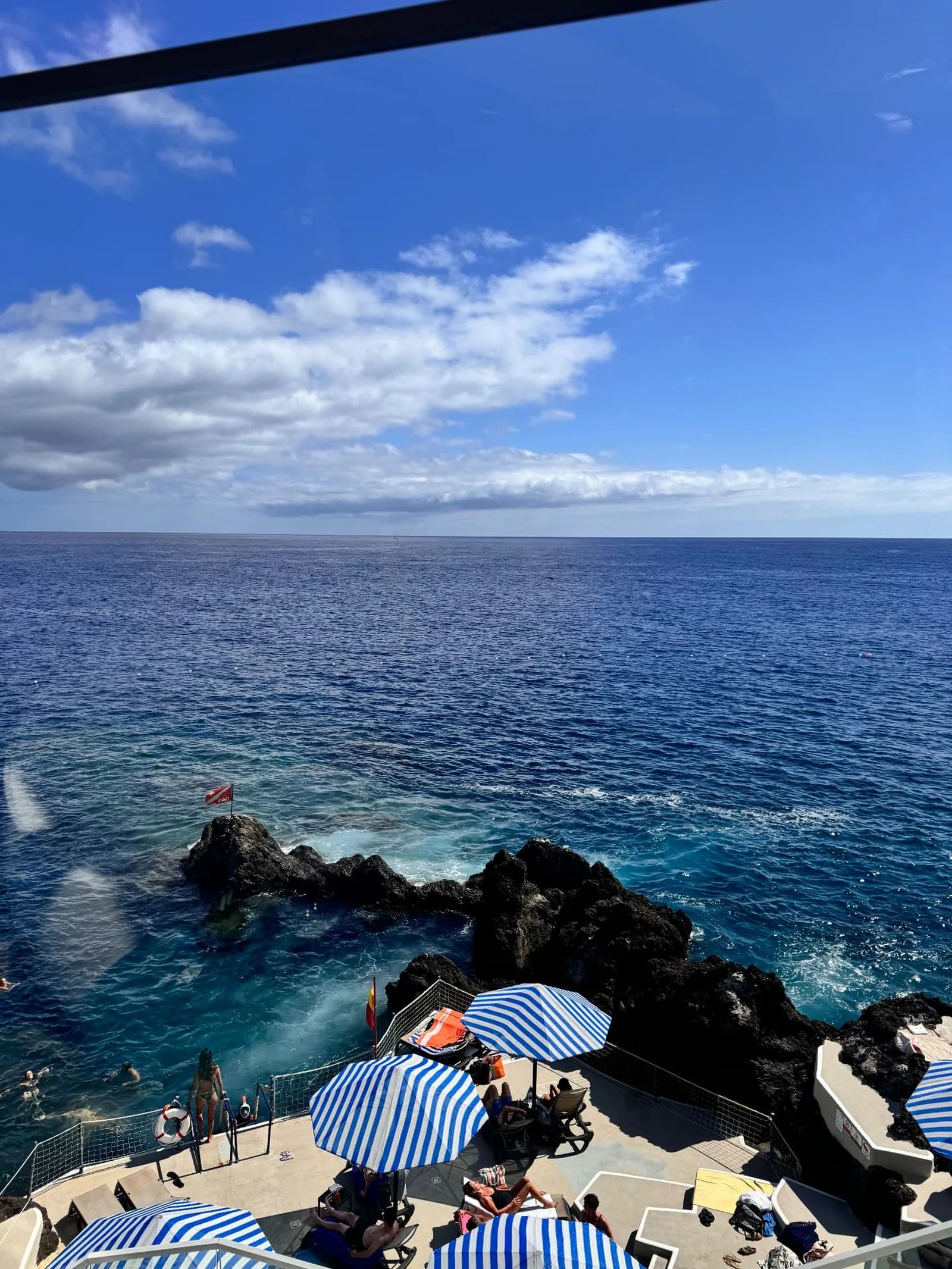 View from Capoeira Lounge over the Caniço de Baixo sea platform, with blue-and-white striped parasols, volcanic rocks, swimmers and a diving flag