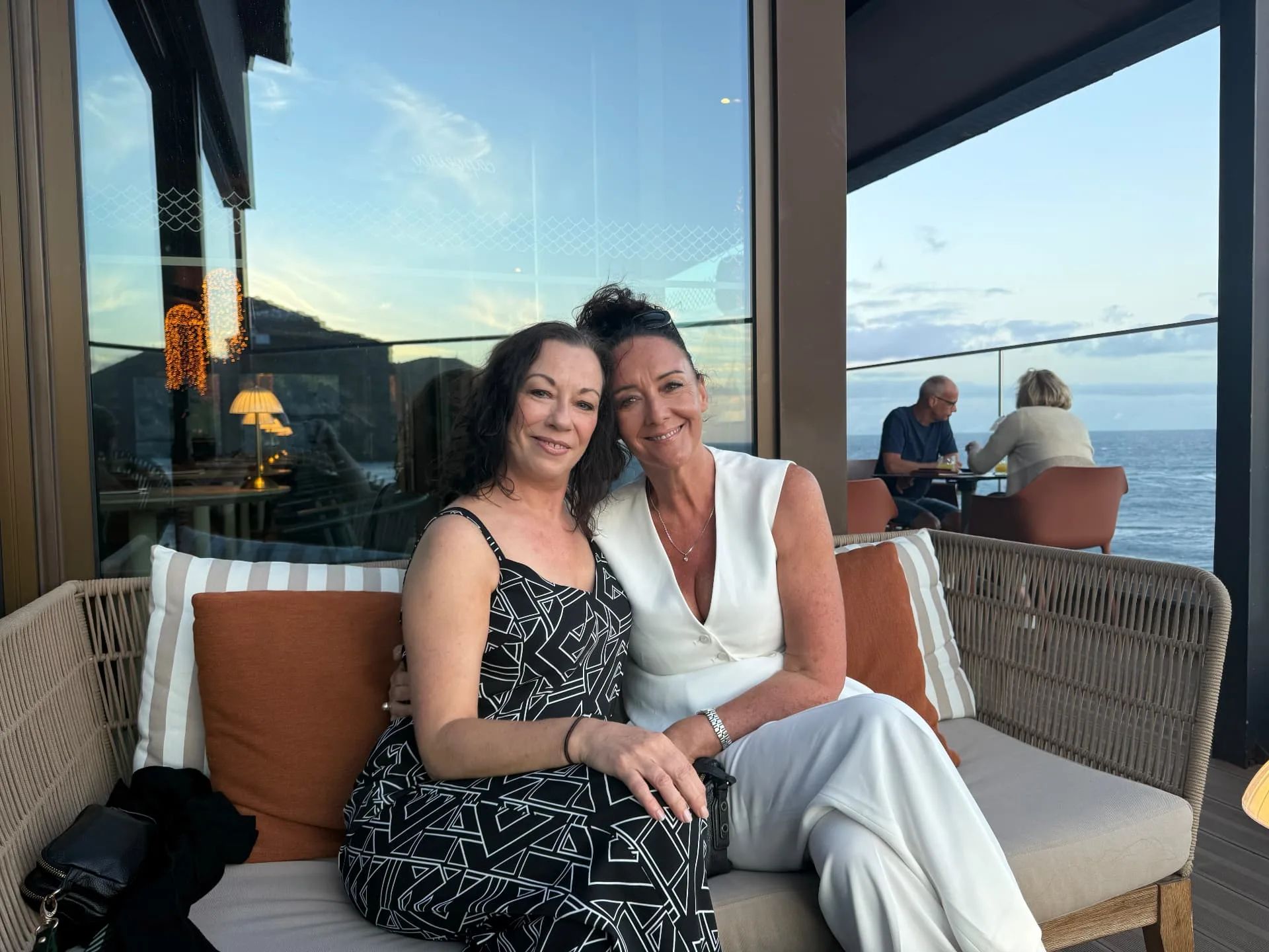 Dois convidados sentados num sofá de vime no Capoeira Lounge, no Caniço de Baixo, ao pôr-do-sol, com janelas do chão ao teto e o Atlântico às suas costas.