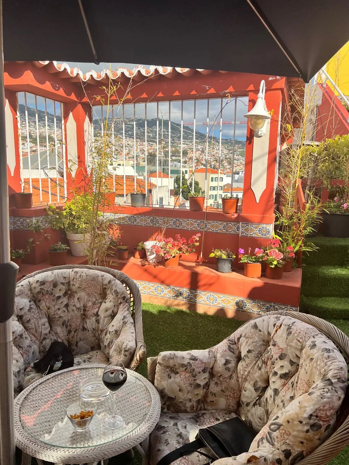 Cadeiras de vime e um copo de vinho tinto no terraço do Flamingo Cocktail Bar, no Se Boutique Hotel, no Funchal, com paredes de terracota, vasos de flores e vistas sobre os telhados da cidade até às colinas