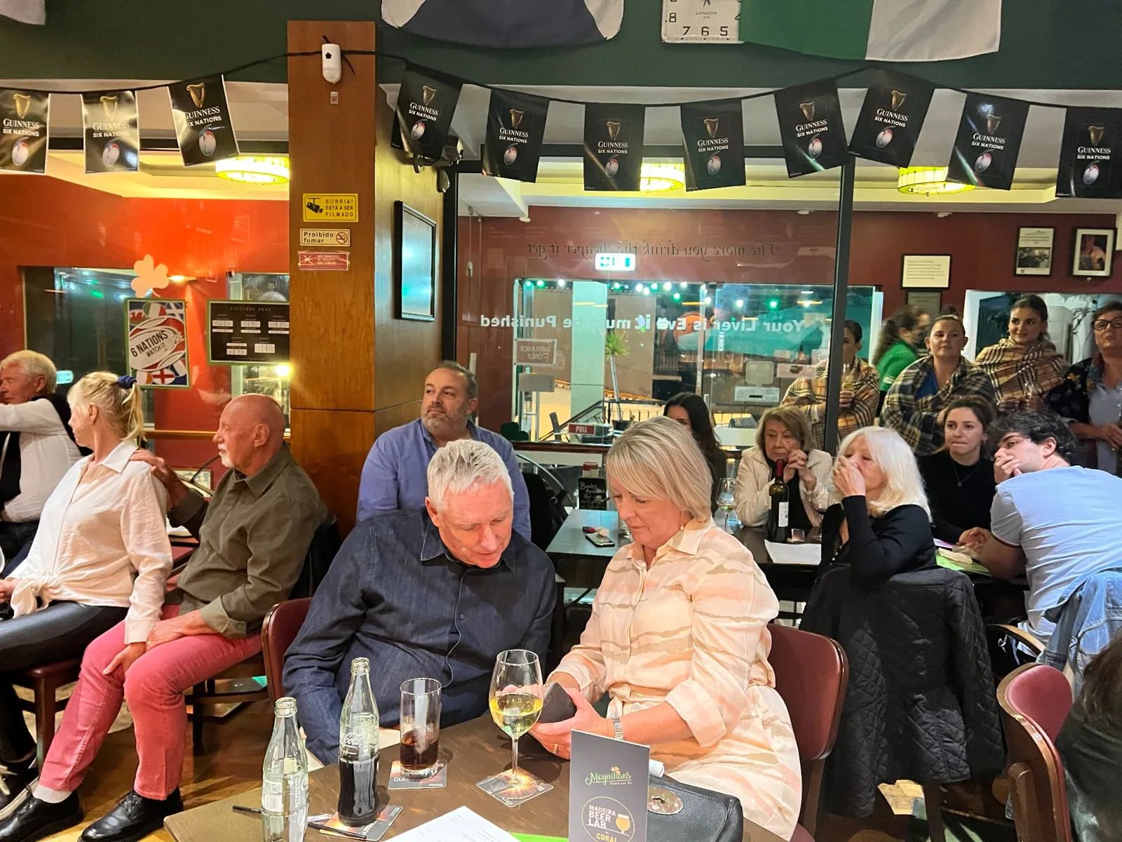Convidados sentados na sala de jantar revestida a painéis de madeira do Moynihans Irish Bar, no Funchal, sob bandeirinhas da Guinness Six Nations