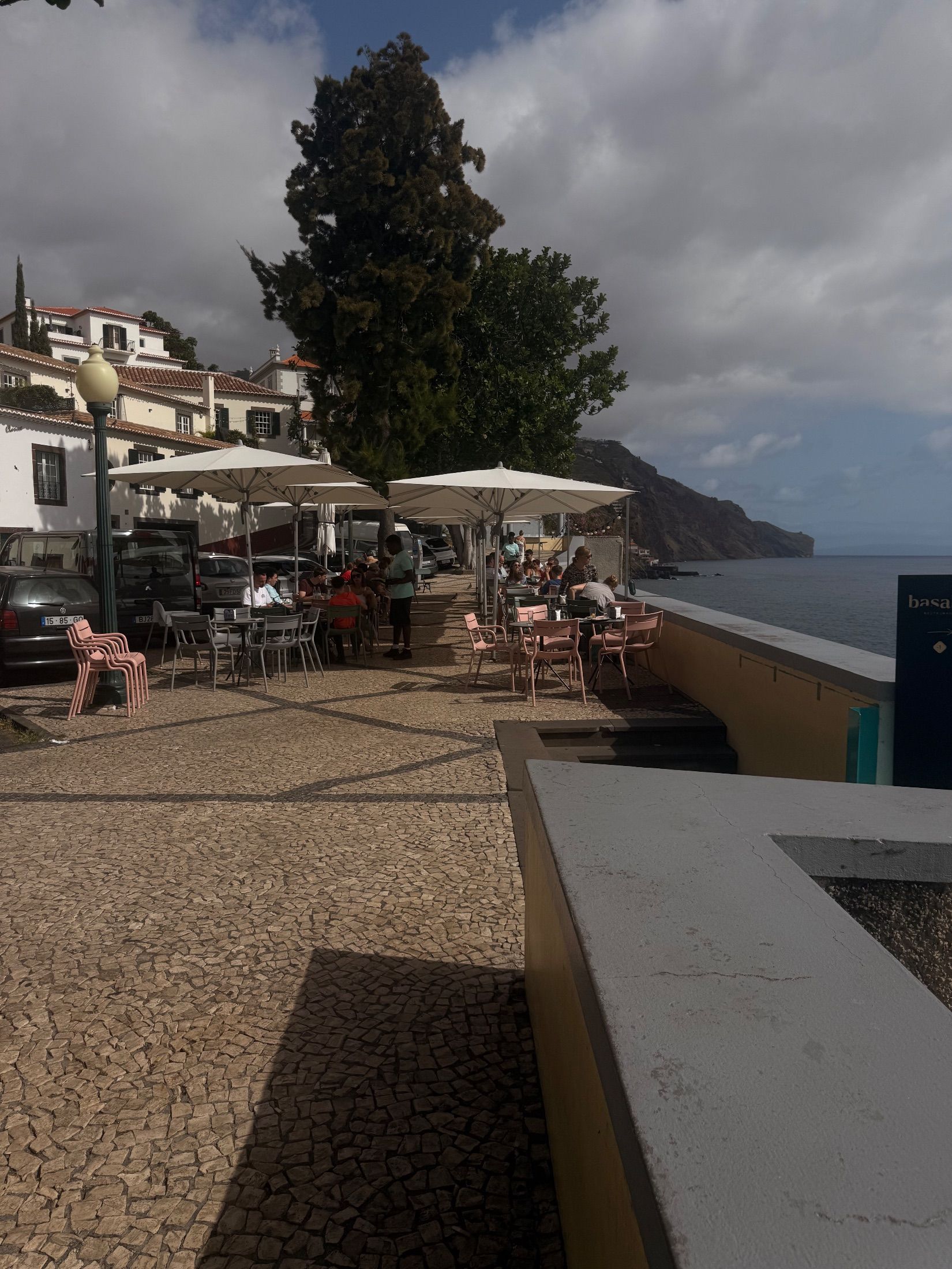 Esplanada à beira-mar com guarda-sóis, clientes sentados e calçada com padrões