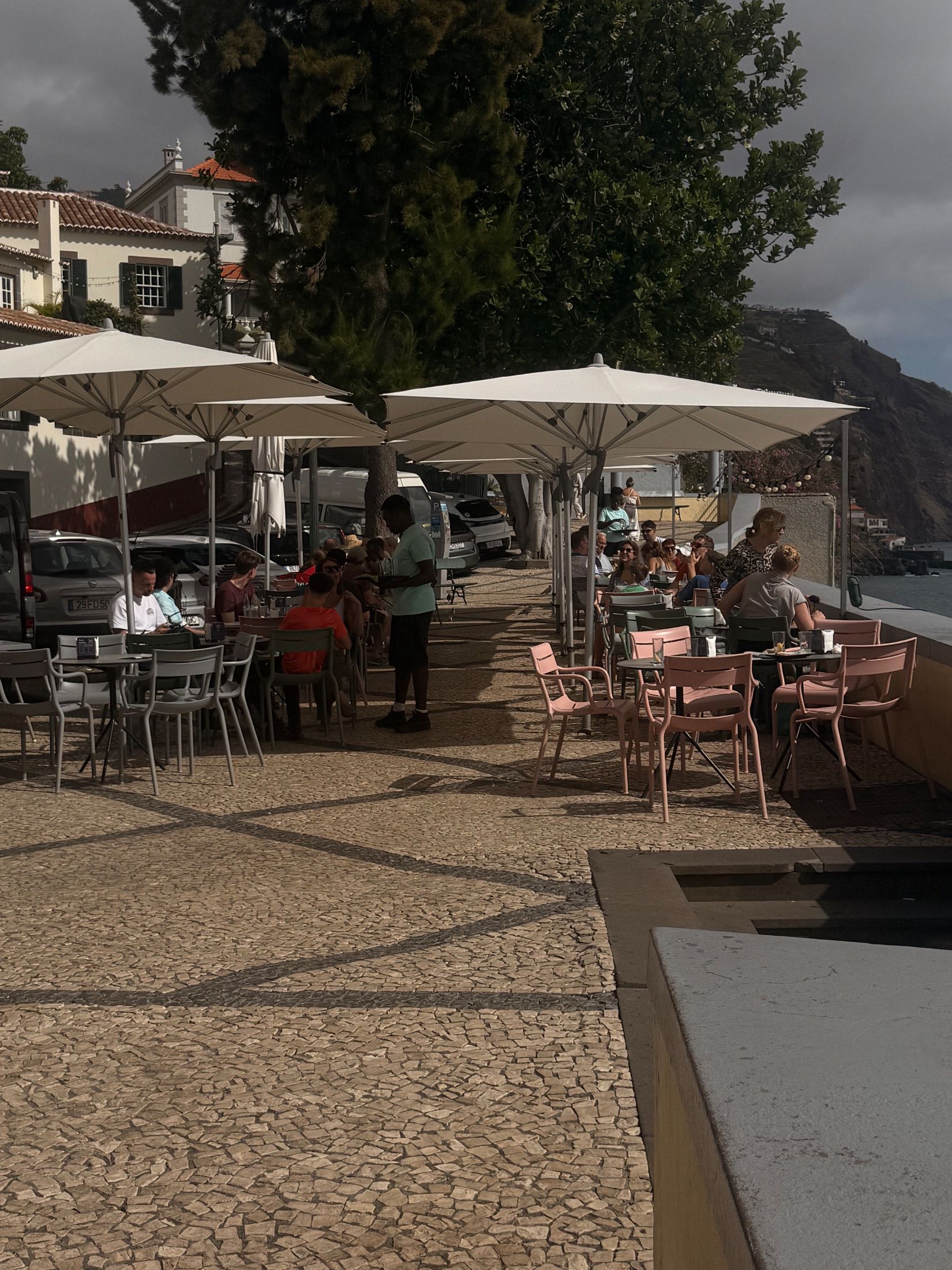 Clientes sentados sob guarda-sóis brancos numa esplanada calcetada com vista para o mar