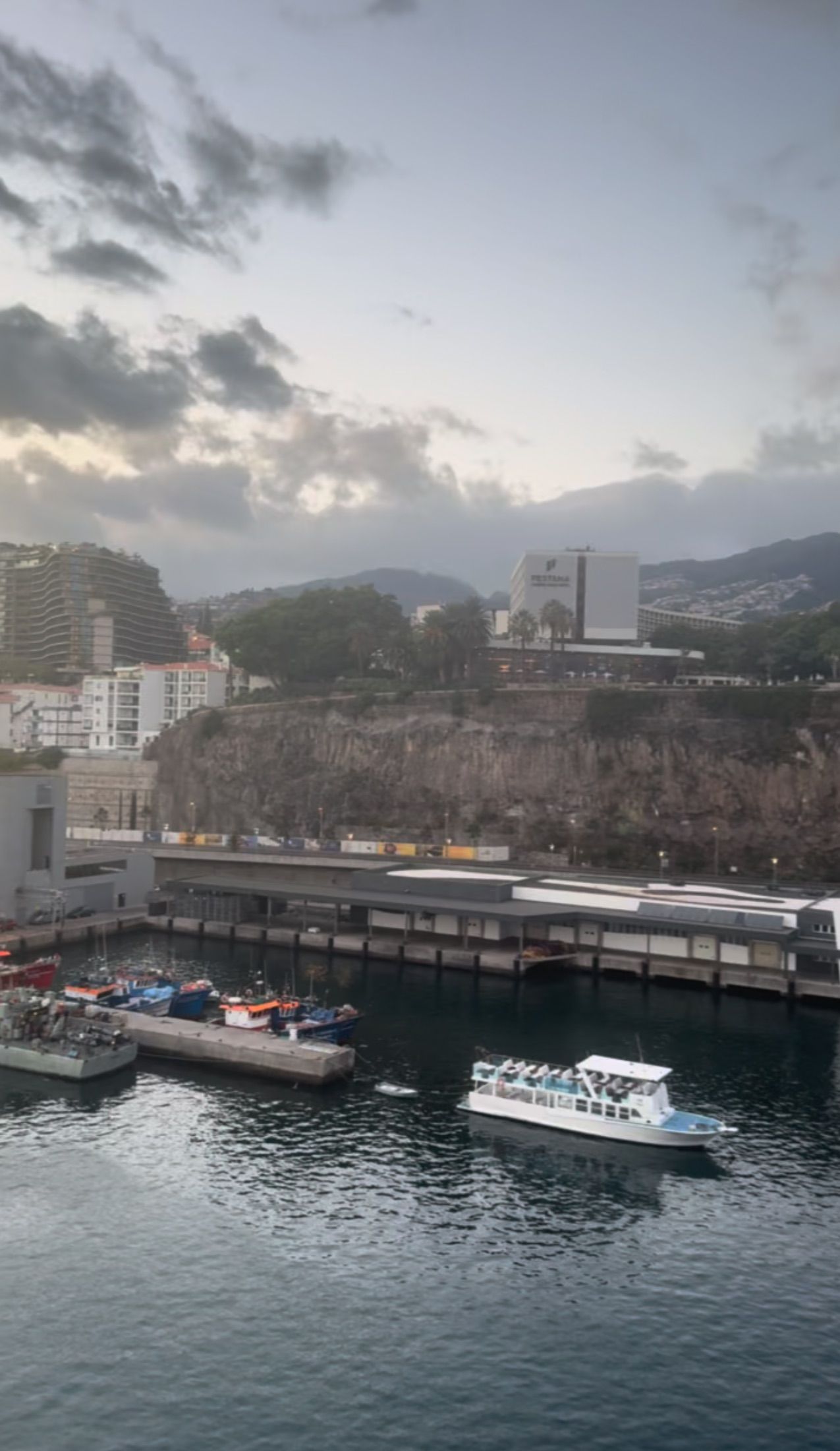 Barcos num porto sob uma escarpa rochosa, edifícios à beira-mar e casas na encosta