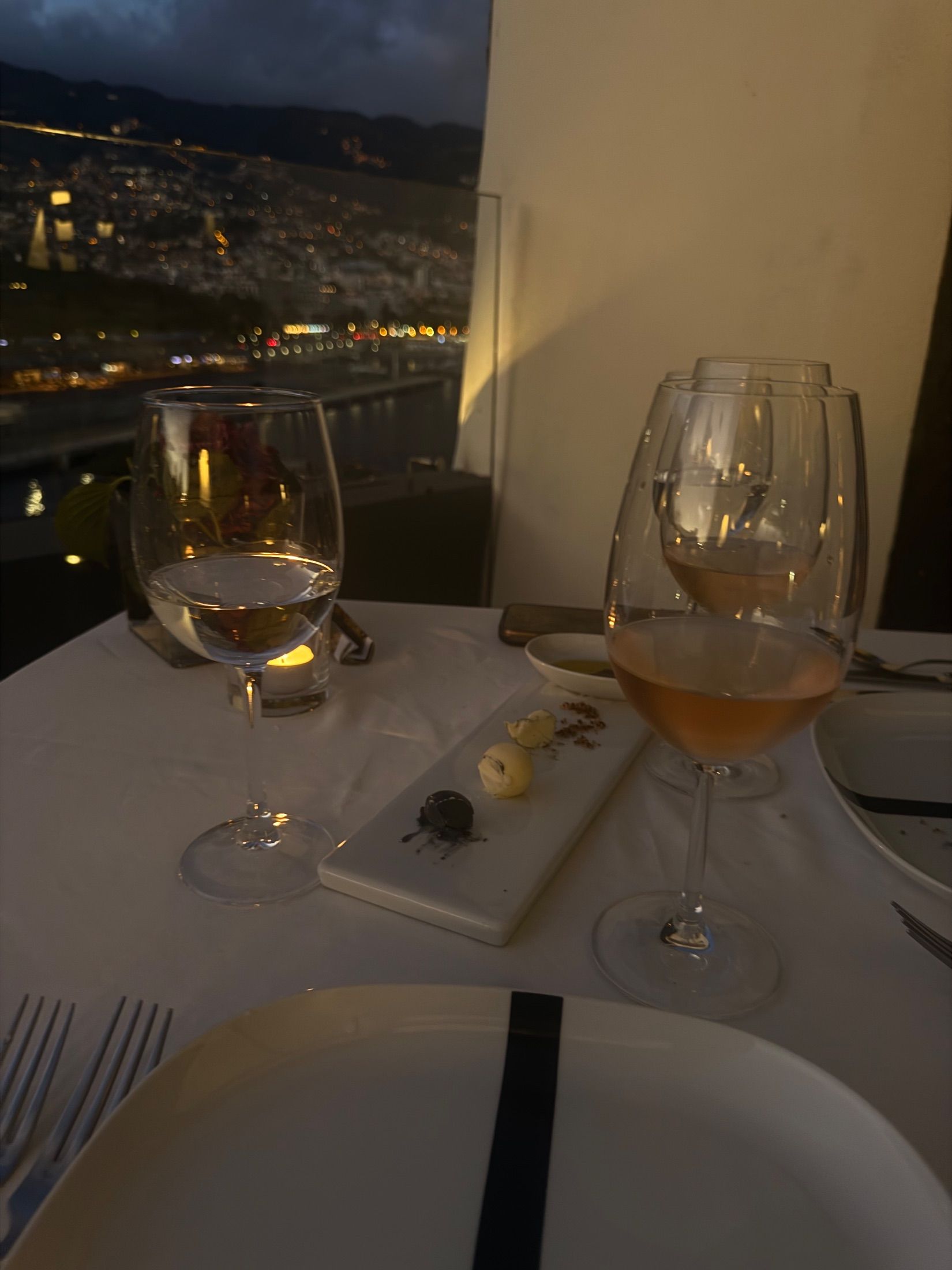 Copos de pé e pratos numa mesa à luz de vela sobre as luzes da cidade