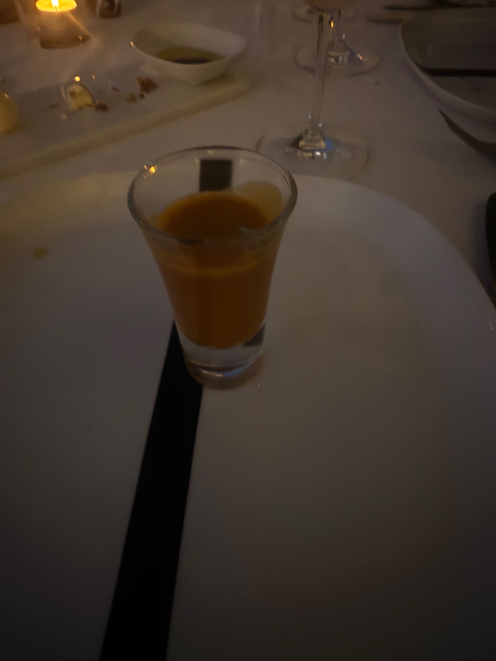 Small glass of orange liquid on a white plate beside stemmed glasses