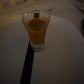 Small glass of orange liquid on a white plate beside stemmed glasses