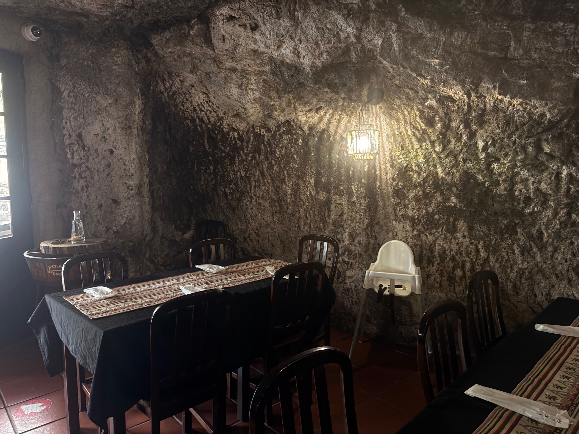 Mesas e cadeiras de madeira junto a uma parede rochosa com um candeeiro aceso