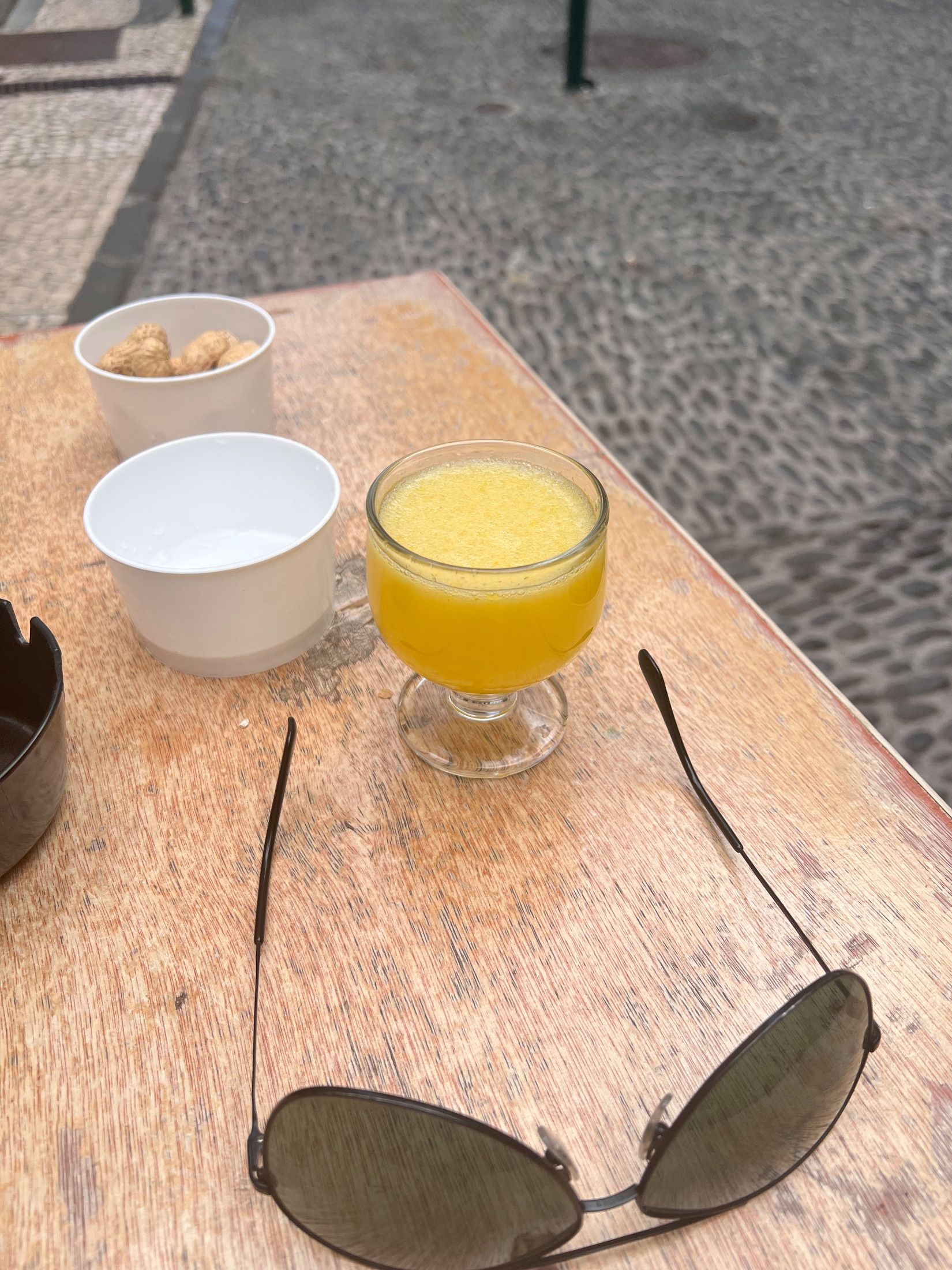 Yellow drink, peanuts and sunglasses on a wooden table beside cobbled paving