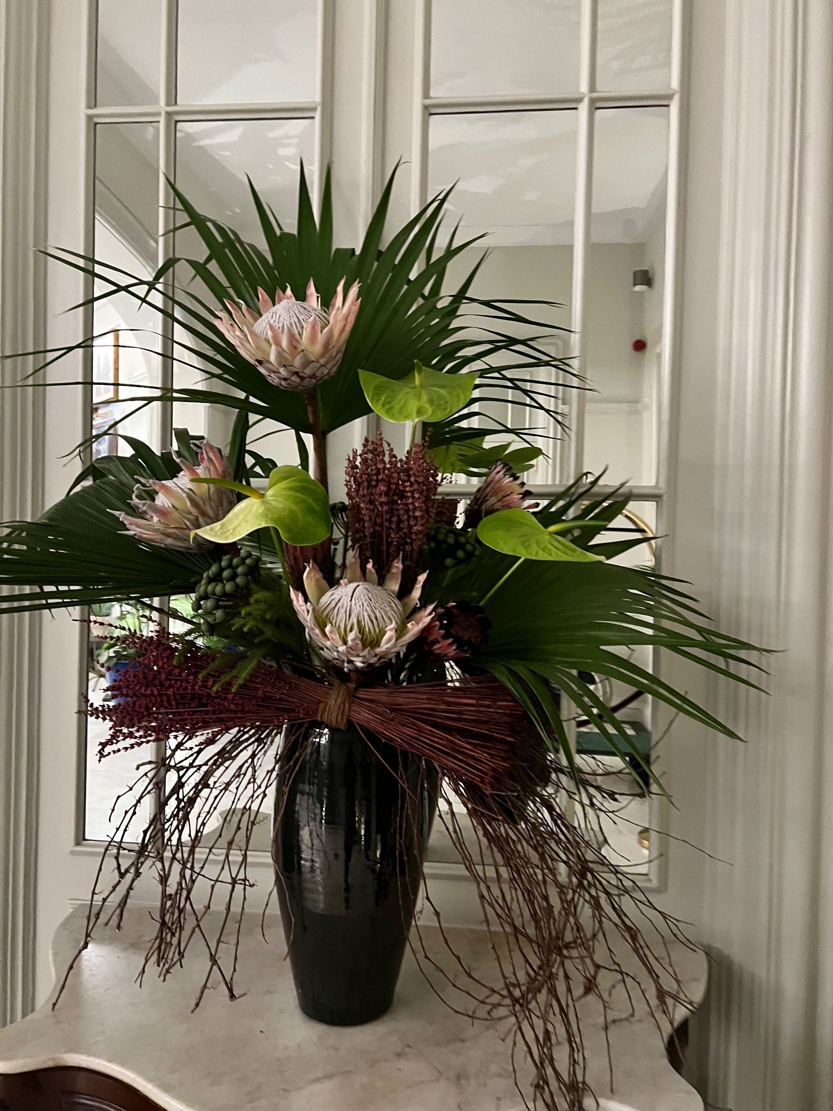 Proteas, green anthuriums and palm leaves arranged in a black vase before a mirror