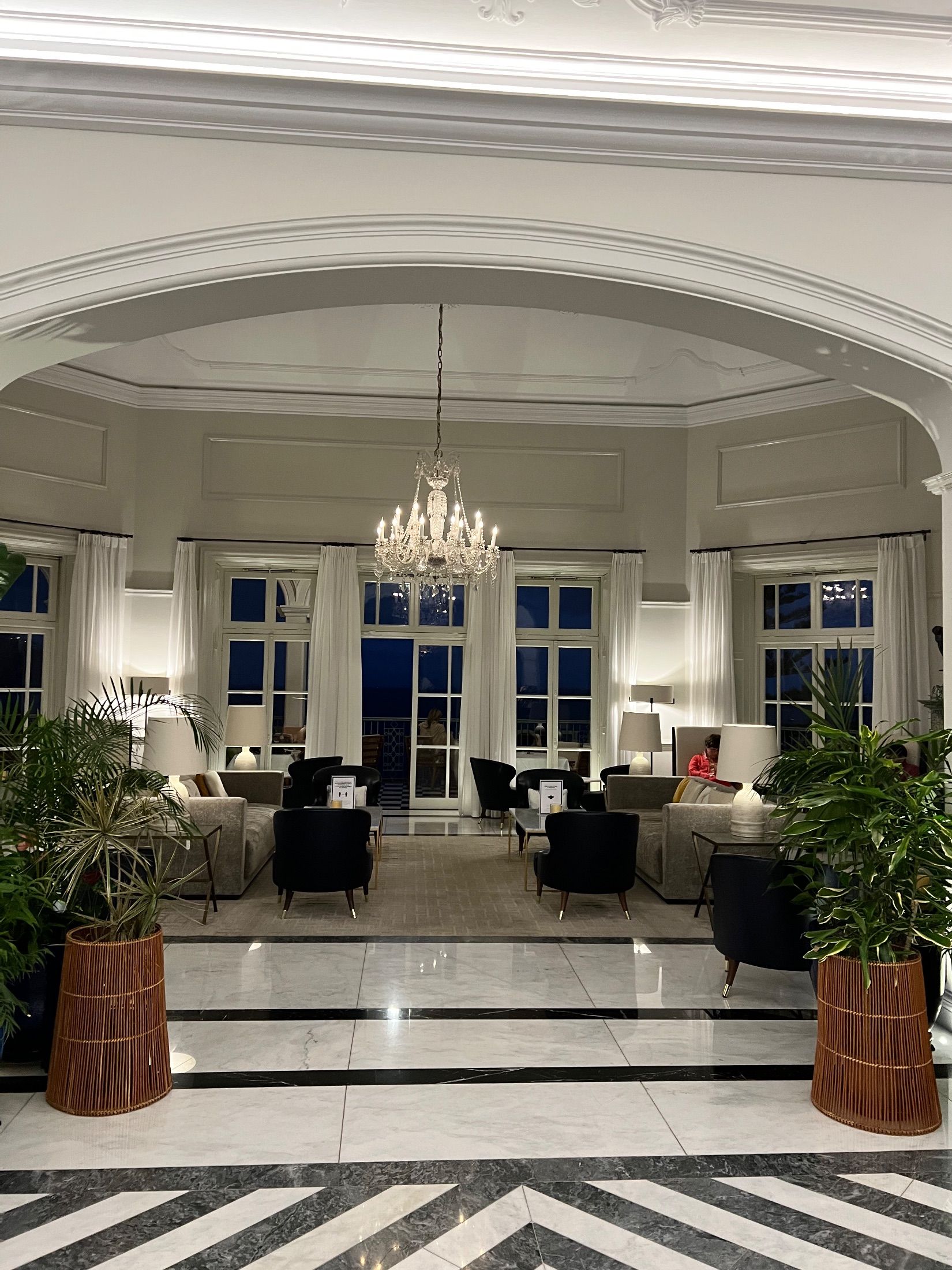Lounge with a chandelier, sofas, black armchairs and potted plants beneath a wide arch