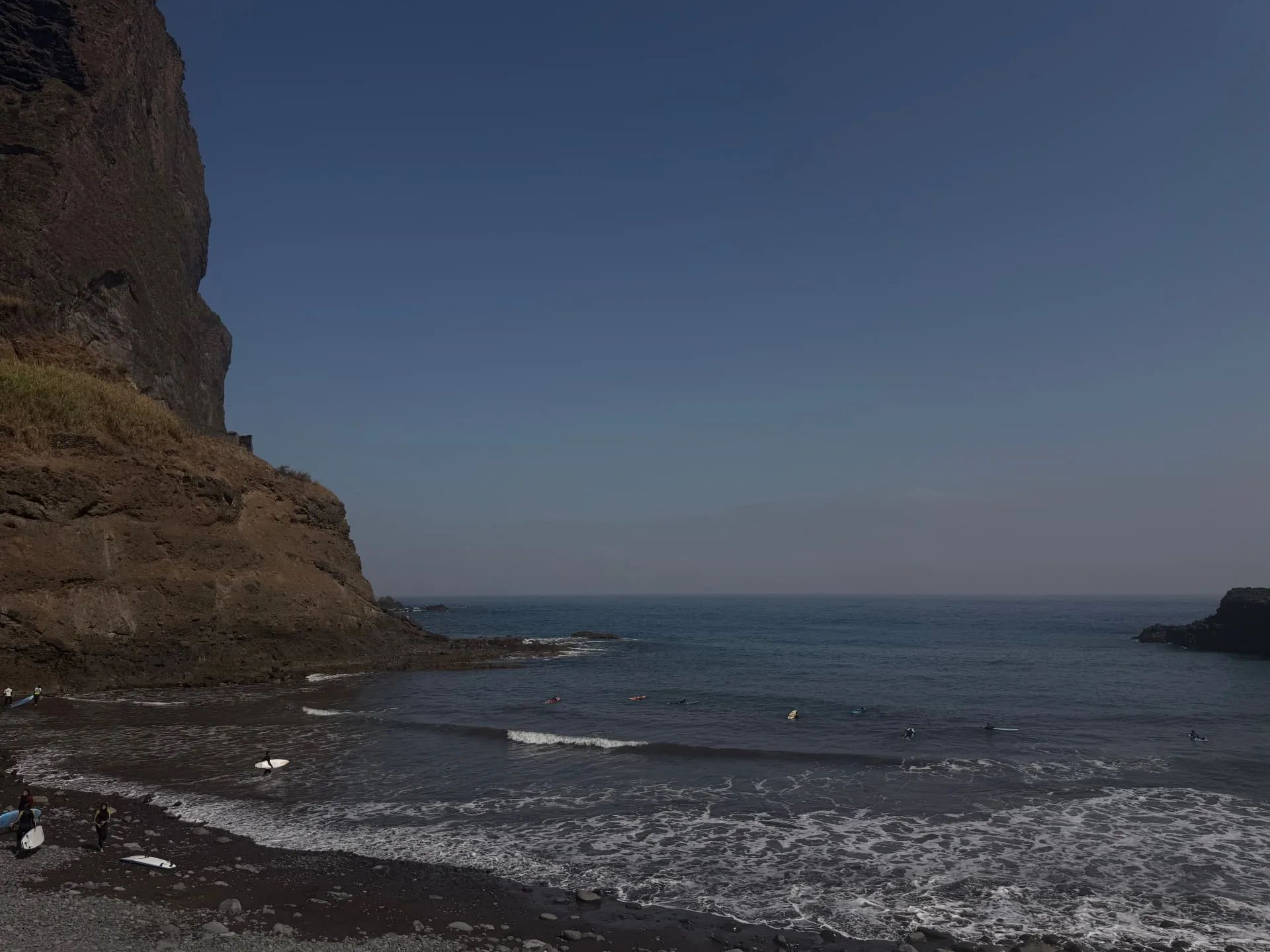 Surfers spread across a small incoming wave at Praia da Alagoa beneath the dark volcanic headland.