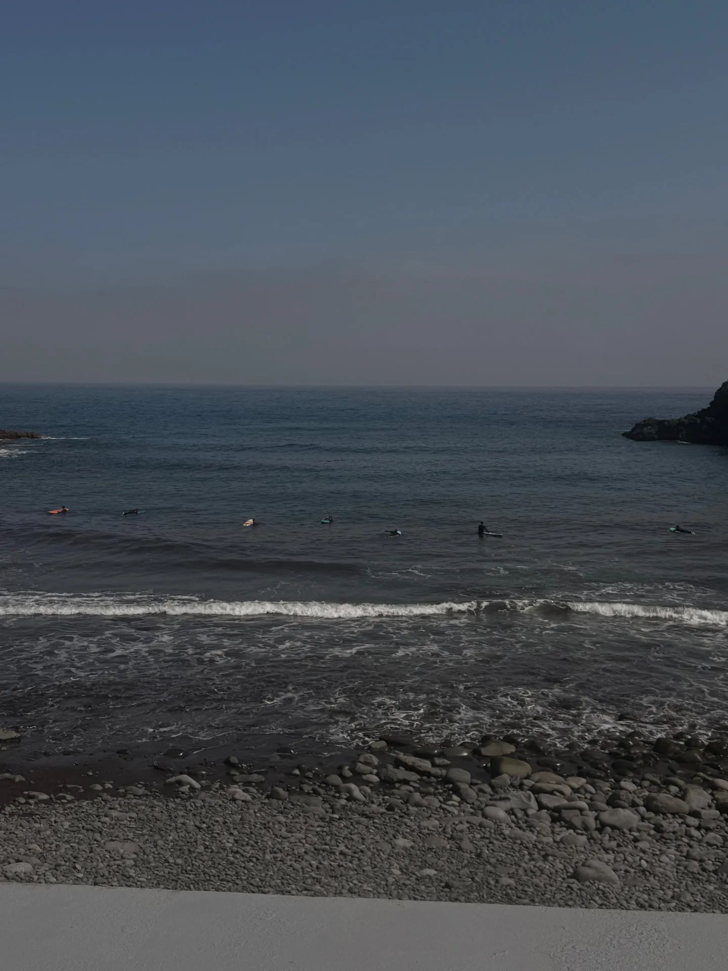 Surfers sitting beyond the shore break at Praia da Alagoa, seen across the pebble beach.