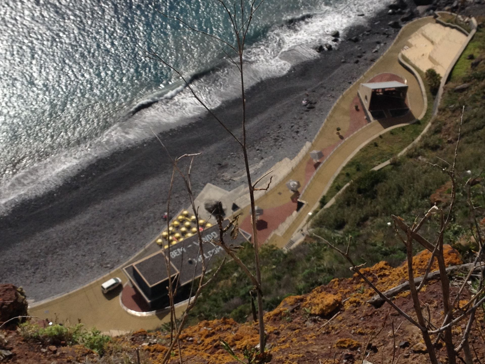 View from the top of the Cristo Rei cable car looking down at Praia do Garajau, the pebble beach, the sea and the small cafe with yellow umbrellas.