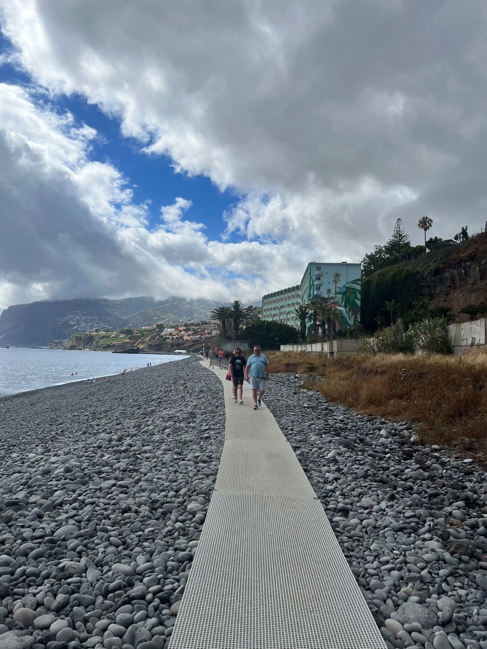 People walking along a narrow walkway across a pebble beach beside the sea