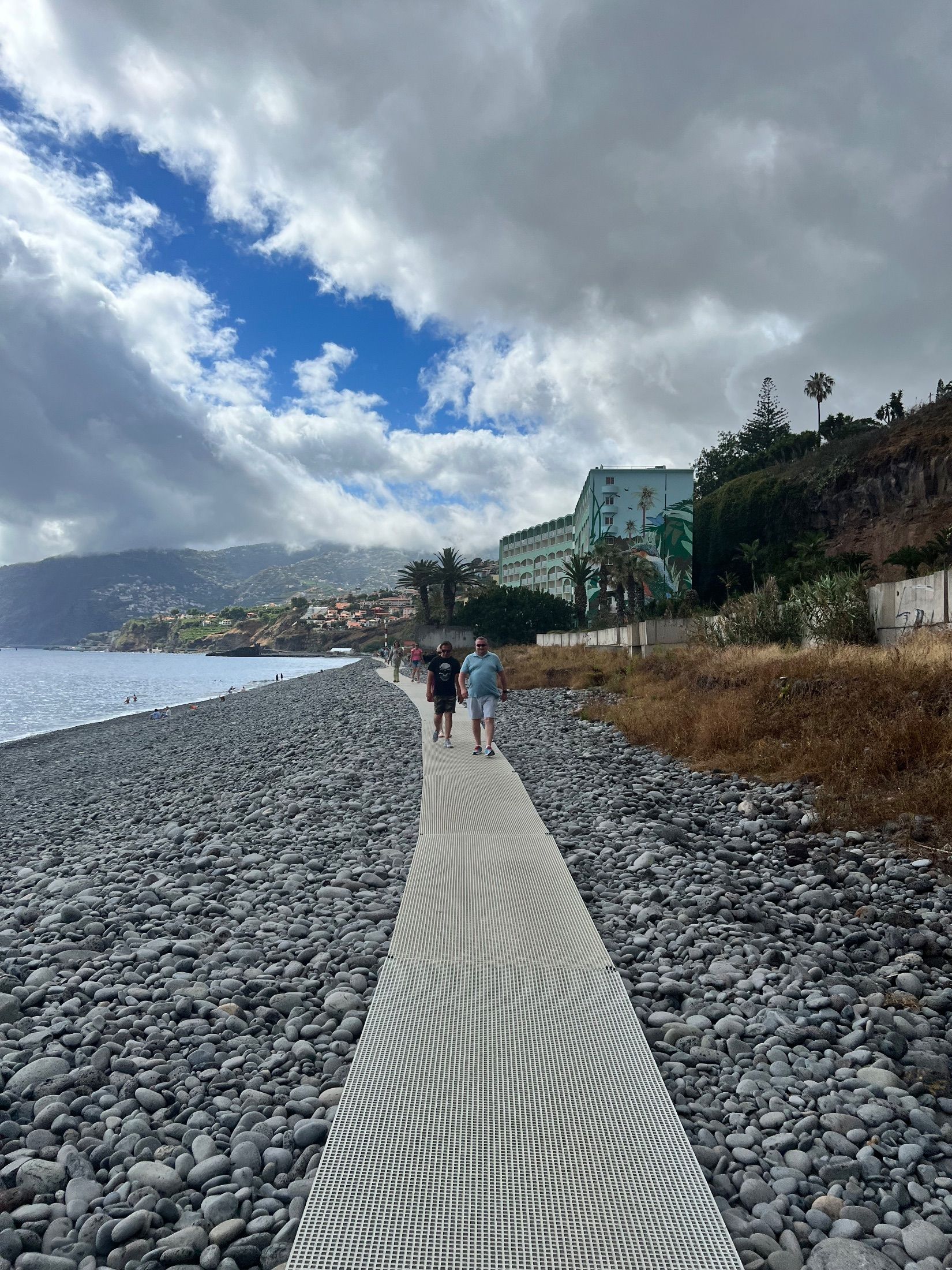 People walking along a narrow walkway across a pebble beach beside the sea