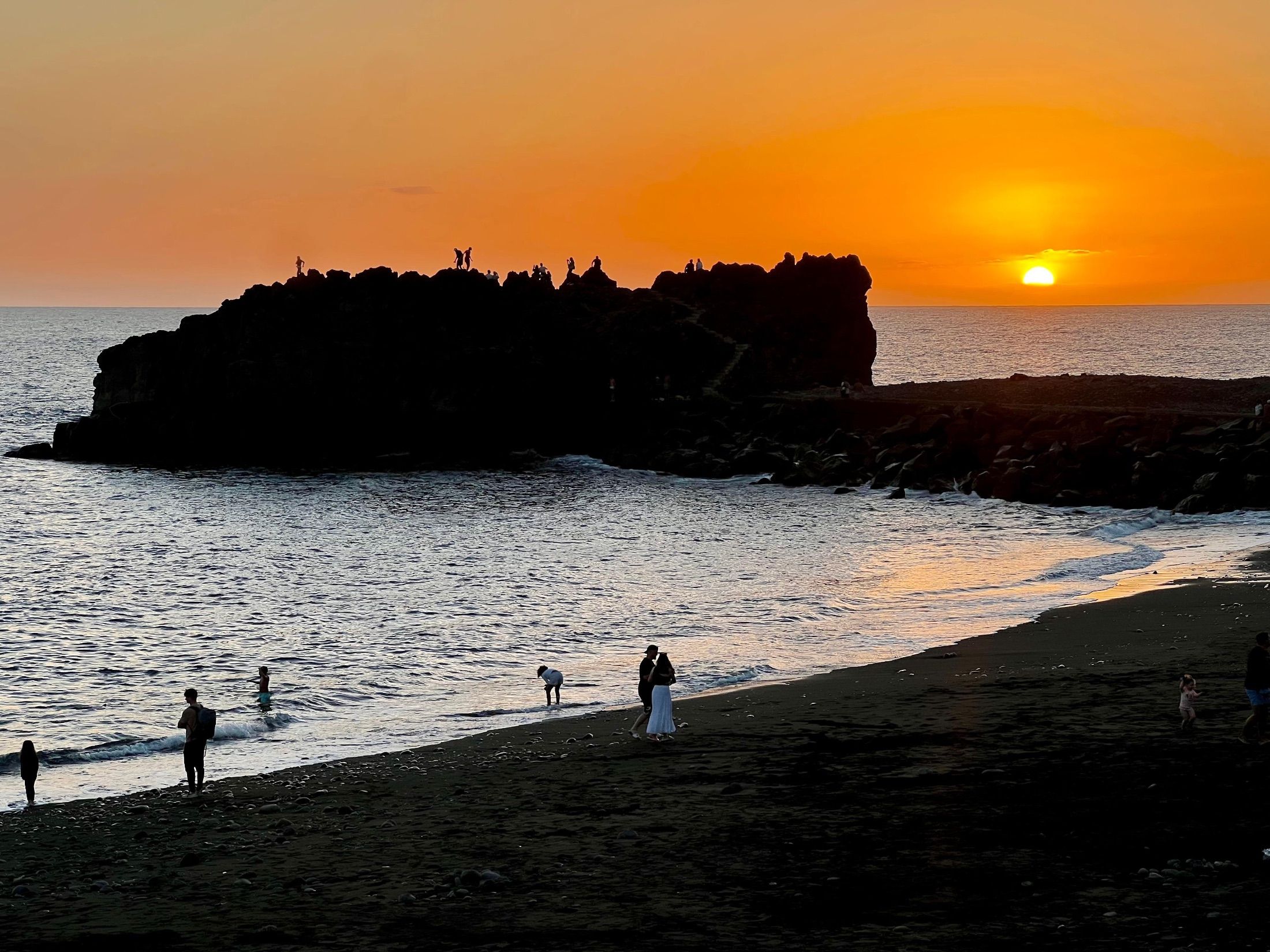 People on a dark sandy beach beneath a rocky outcrop at sunset