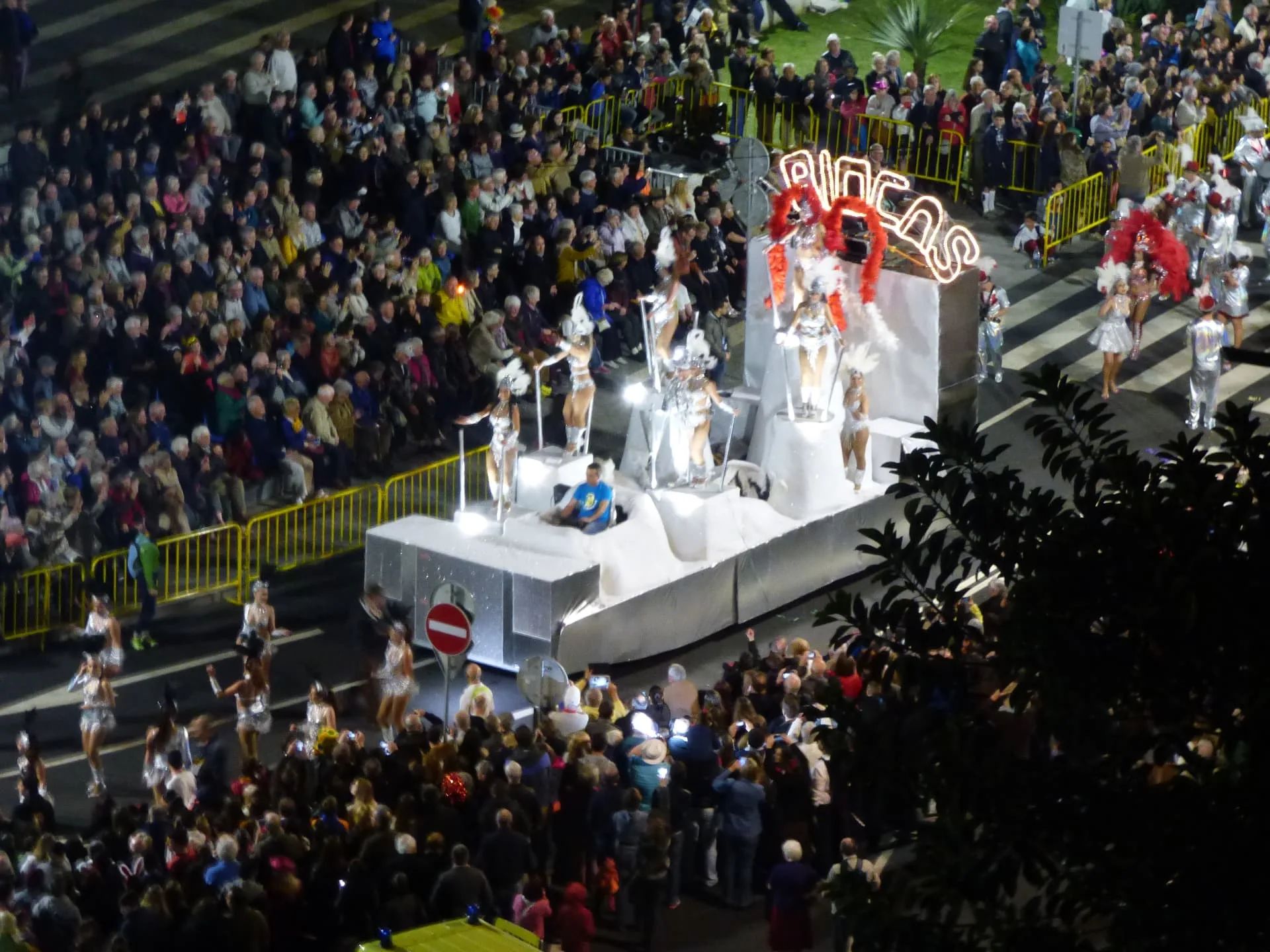 Silver-and-white carnival float with feathered samba dancers passing packed crowds at night during the Madeira Carnival parade in Funchal