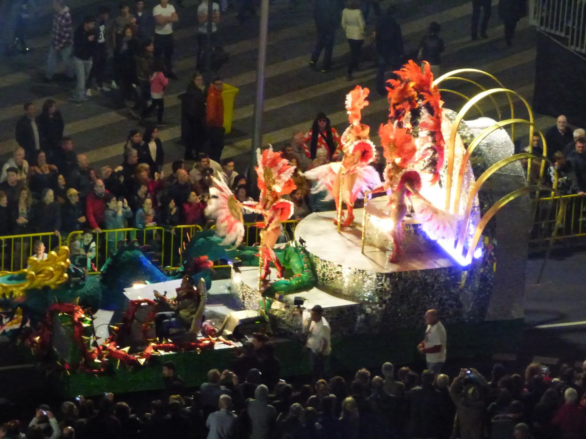 Mirrored carnival float with a green dragon and dancers in red and pink feathers during the Madeira Carnival night parade