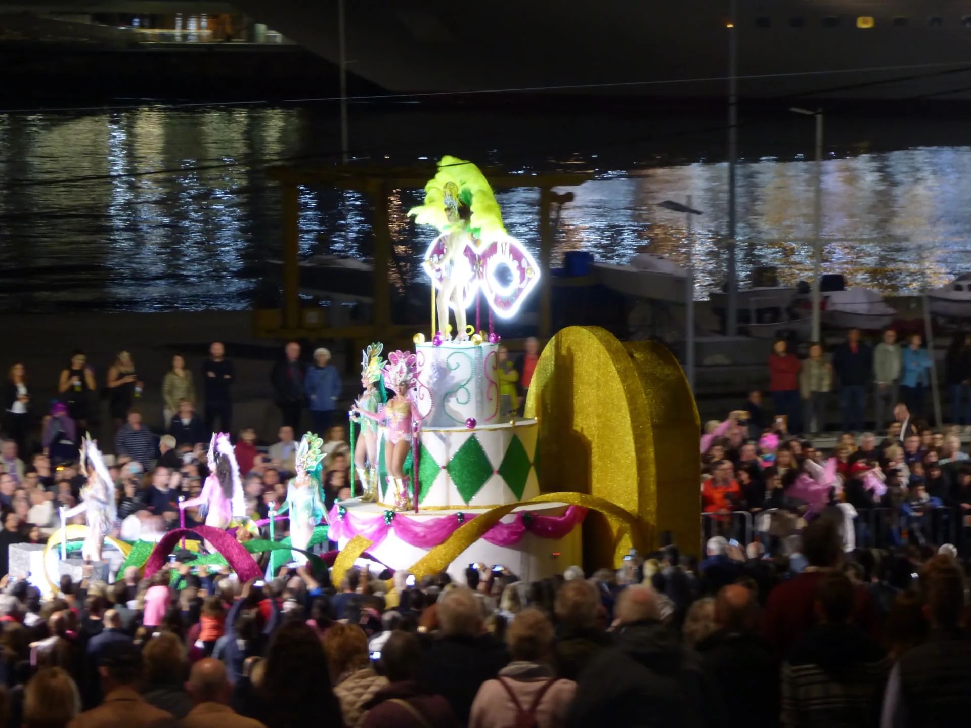 Illuminated cake-shaped carnival float with dancers passing crowds along the Funchal waterfront at night