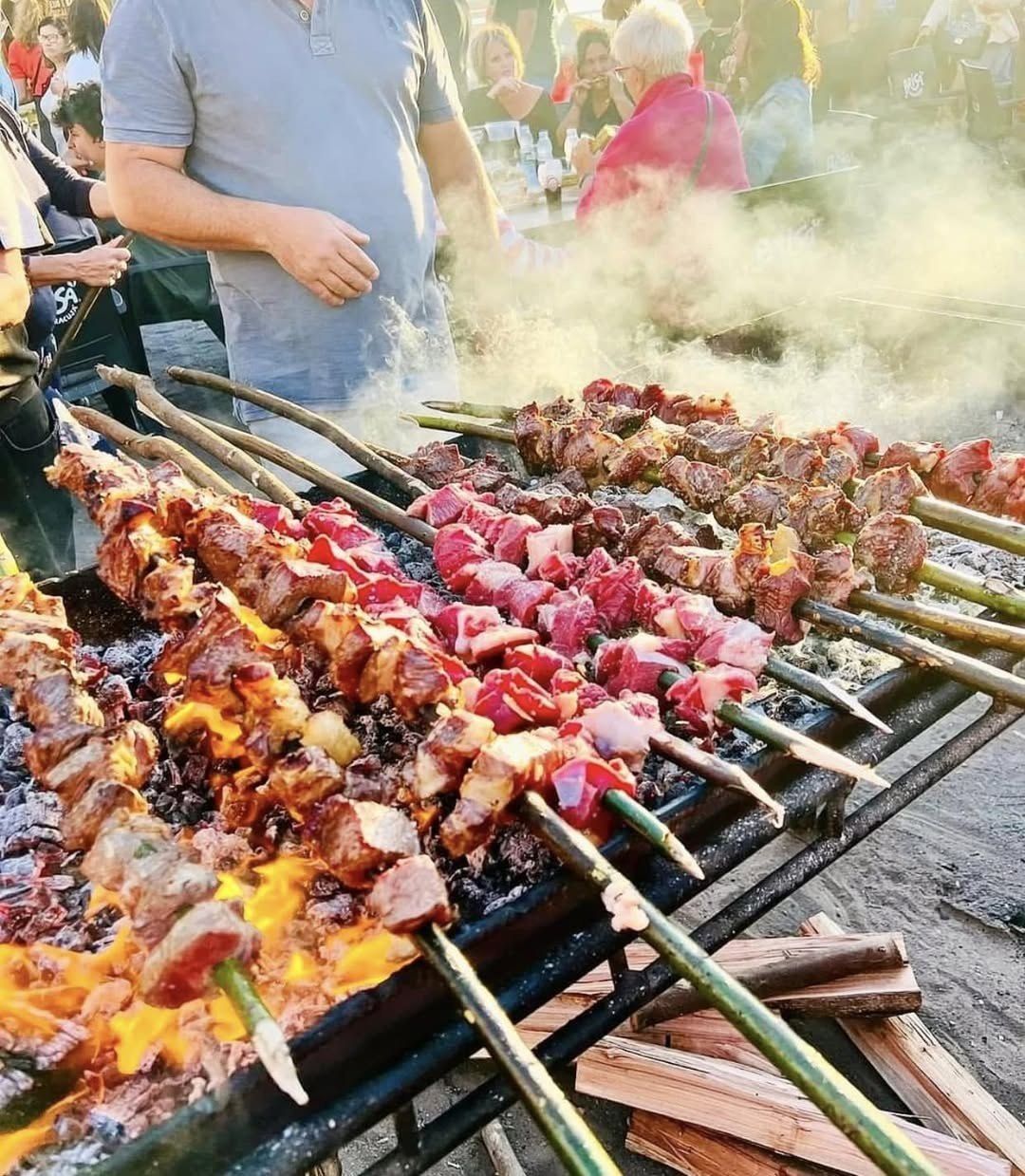 Espetada beef skewers grilling over wood embers on a long charcoal grill at a village arraial festival in Madeira