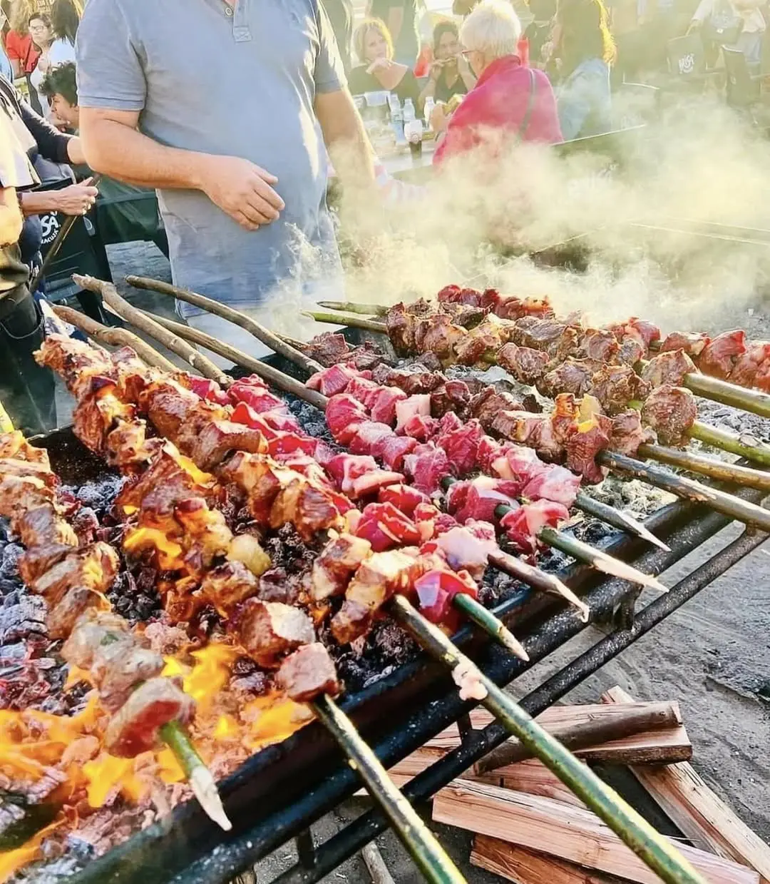 Espetada beef skewers grilling over wood embers on a long charcoal grill at a village arraial festival in Madeira