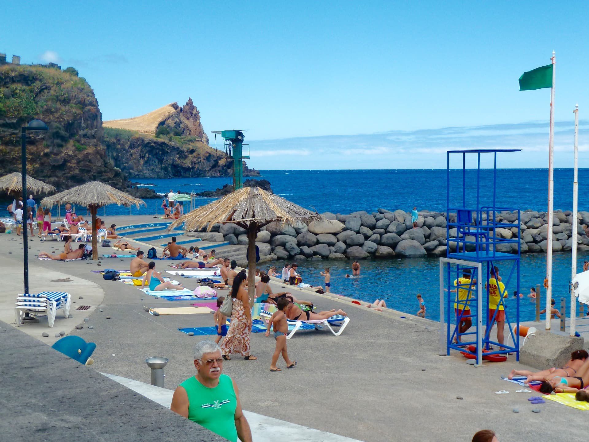 Busy seaside swimming complex on Madeira's rocky shoreline with sun umbrellas.
