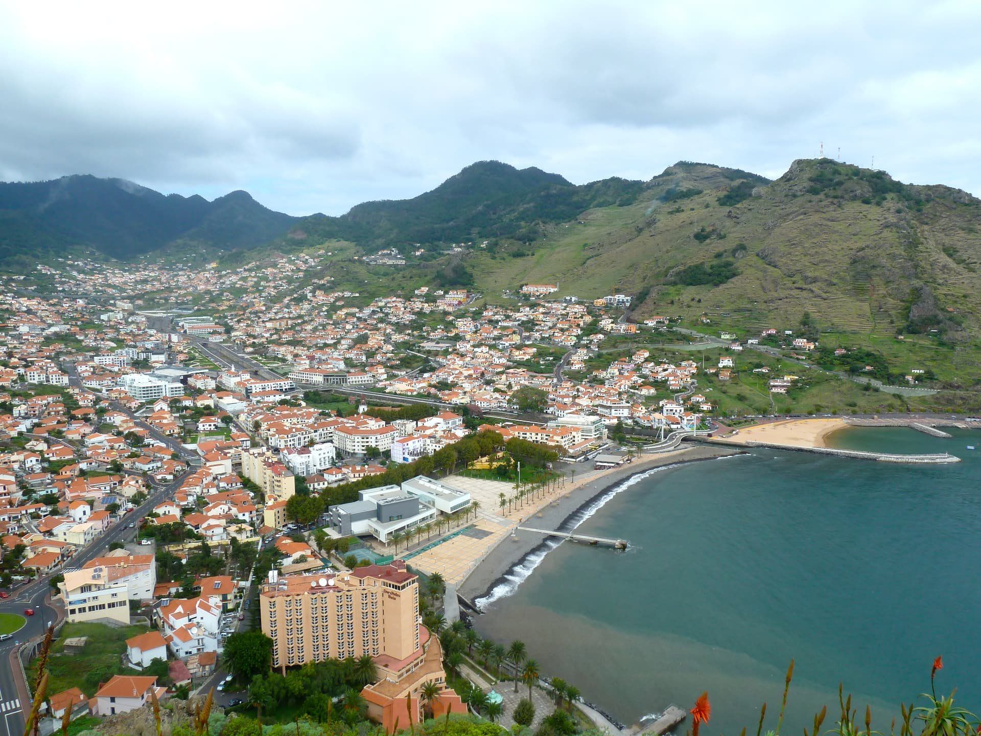 Aerial view over a Madeira coastal town with red roofs, hills and the Atlantic.