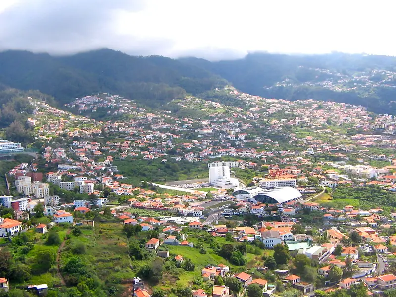 Wide view of a densely built Madeira hillside town below cloudy mountains.