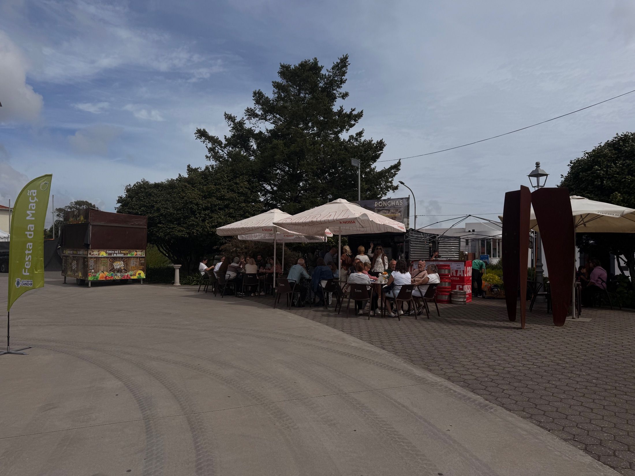 Pessoas sentadas sob chapéus de sol junto a bancas de bebidas e bandeira da Festa da Maçã