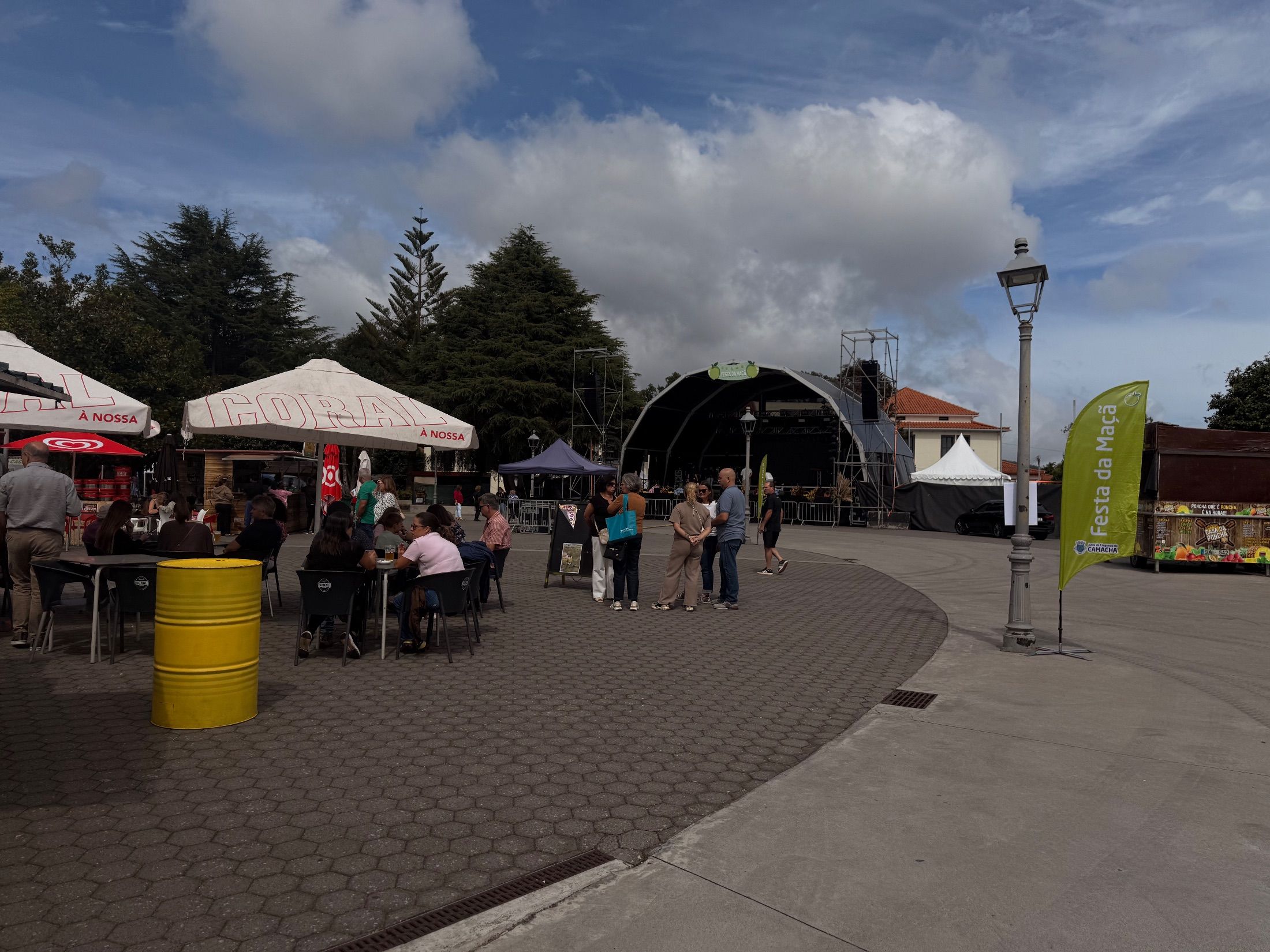 Praça de Camacha - Pessoas em mesas ao ar livre junto a bancas e um palco coberto com bandeiras festivas