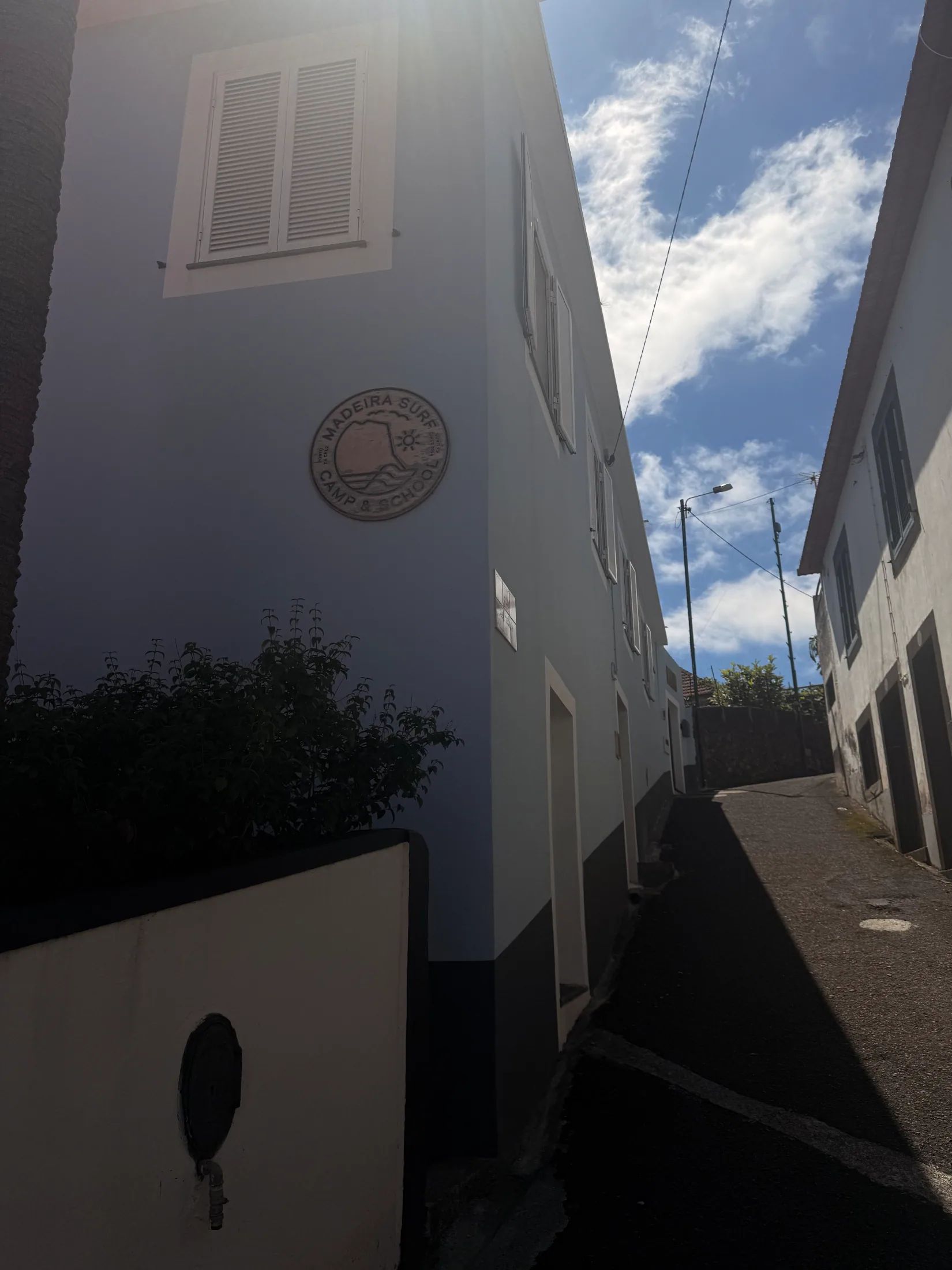 A narrow uphill street between white and blue buildings with a surf camp sign and shutters.