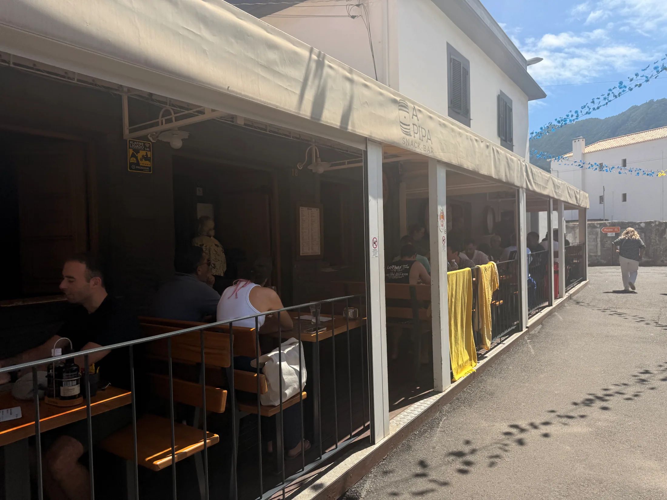 Customers sitting at wooden tables under a large awning at an outdoor terrace of a snack bar.