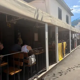 Customers sitting at wooden tables under a large awning at an outdoor terrace of a snack bar.