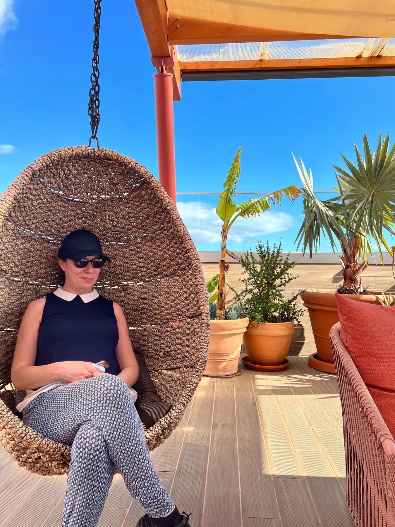 Woman relaxing in a woven hanging chair on the Three House rooftop terrace in Funchal, beside palms and terracotta planters.