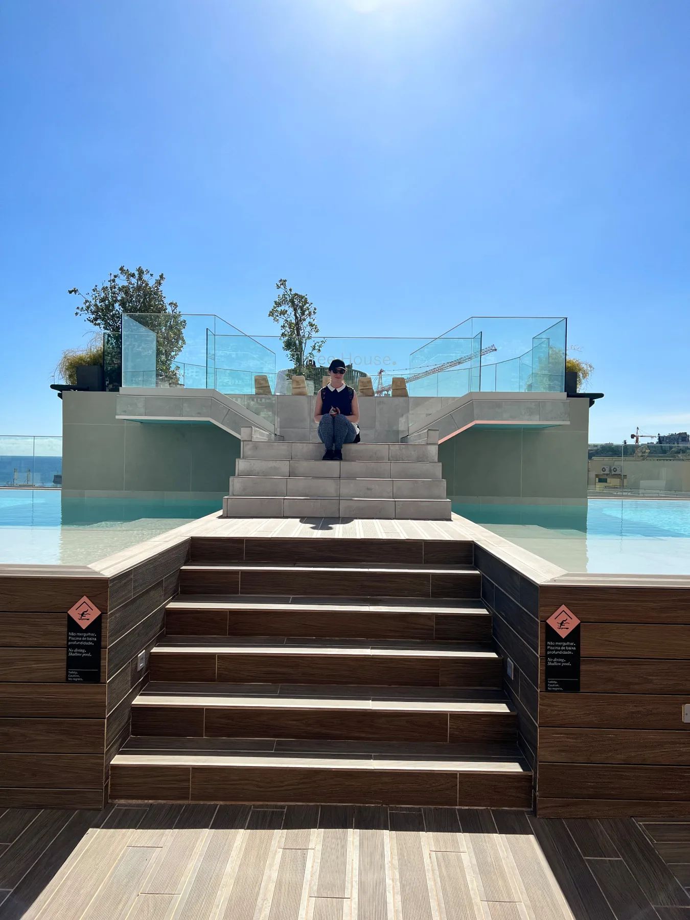 Visitor sitting on the steps beside the Three House rooftop pool in Funchal, with the Atlantic and city skyline behind.