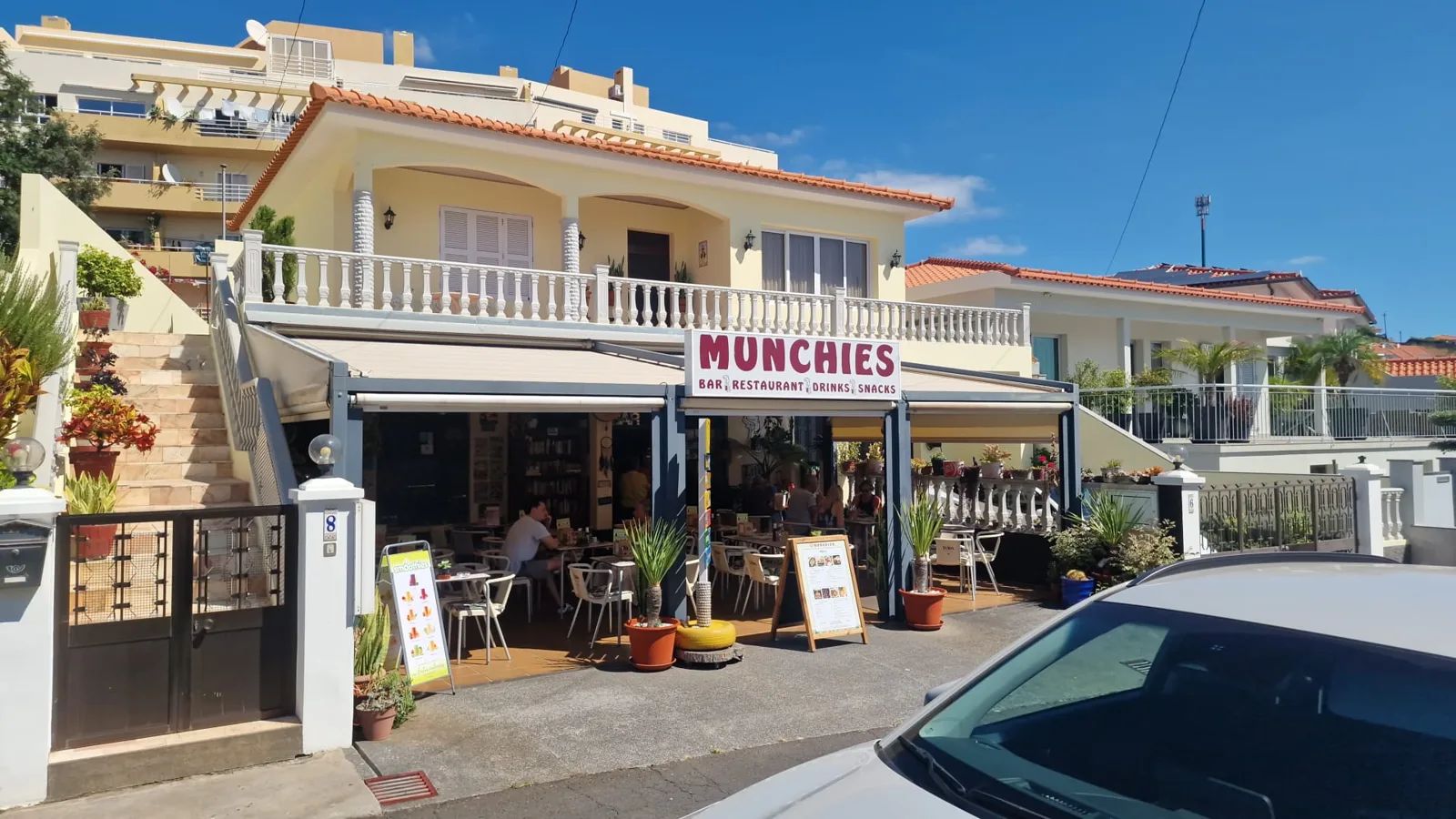 Vista da rua do Munchies Bar Restaurant, no Caniço, na Madeira, com toldos sobre um terraço coberto e clientes sentados no exterior num dia de sol