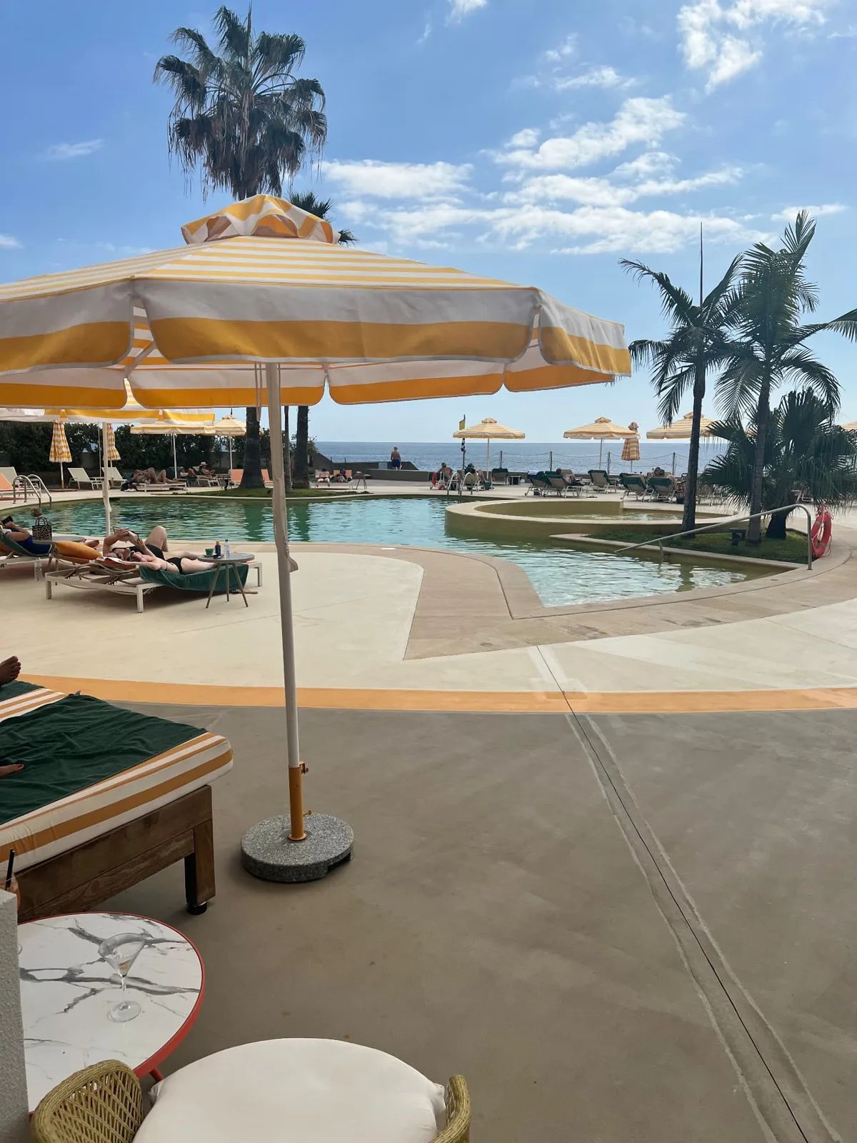 The pool at Recharge restaurant, NEXT Hotel Funchal, with yellow-striped parasols, loungers, palm trees and the Atlantic Ocean in the background