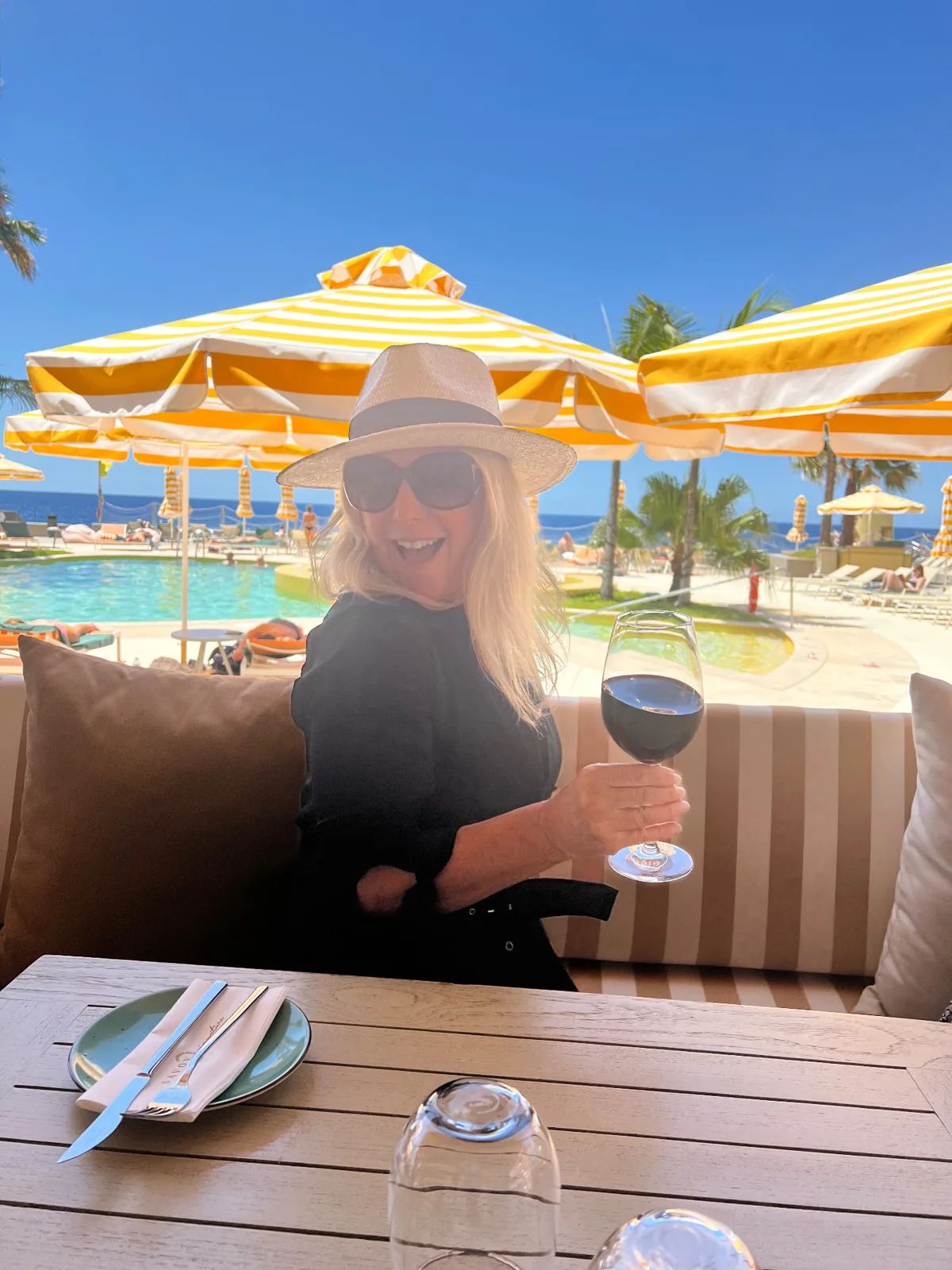 A guest with a glass of red wine on the terrace at Recharge restaurant, NEXT Hotel Funchal, with the pool, yellow-striped parasols and the Atlantic behind