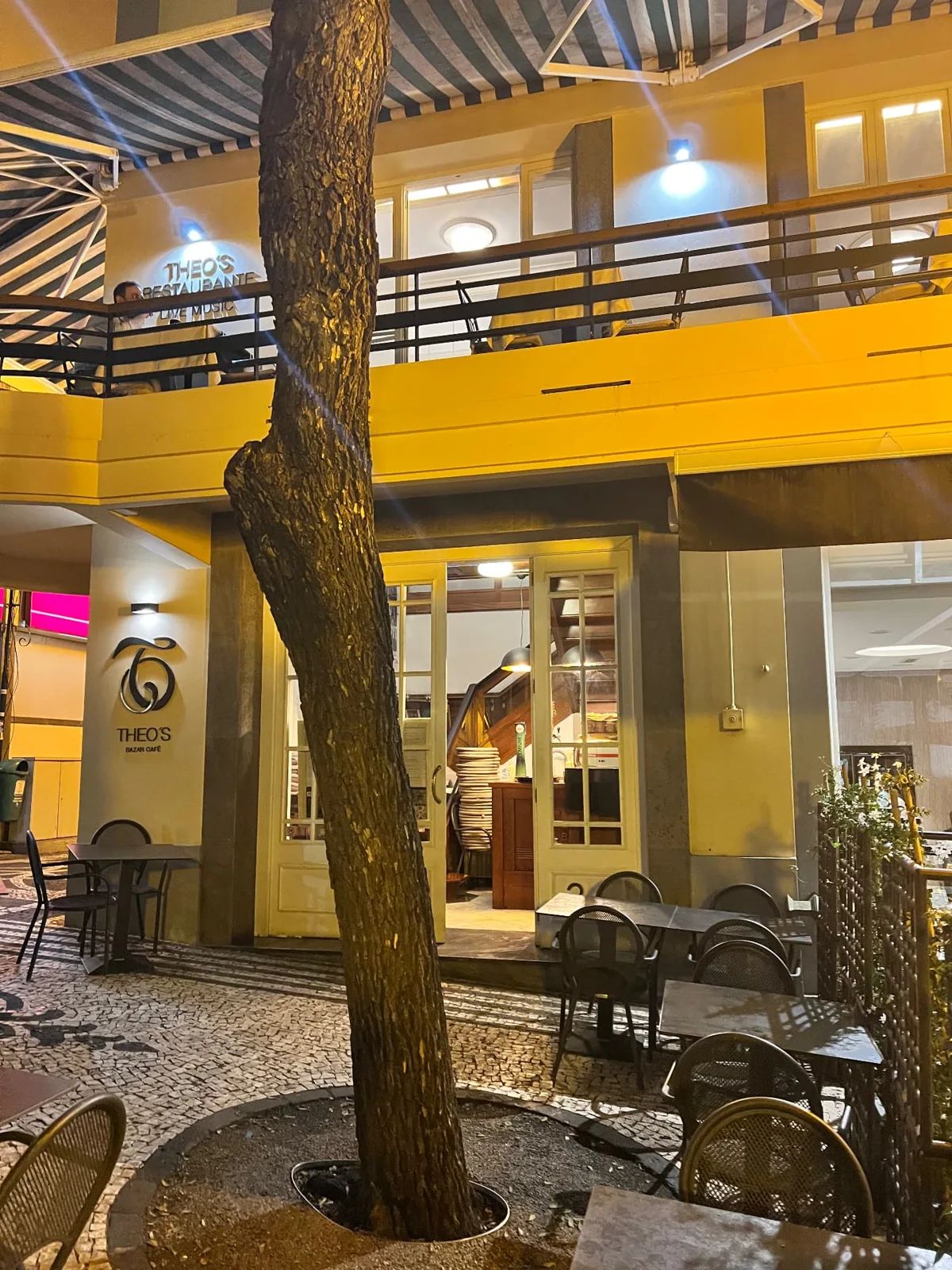 A entrada do Restaurante Theo, no Funchal, à noite, com esplanada de calçada, guarda-sóis às riscas e a sala de jantar do andar de cima iluminada