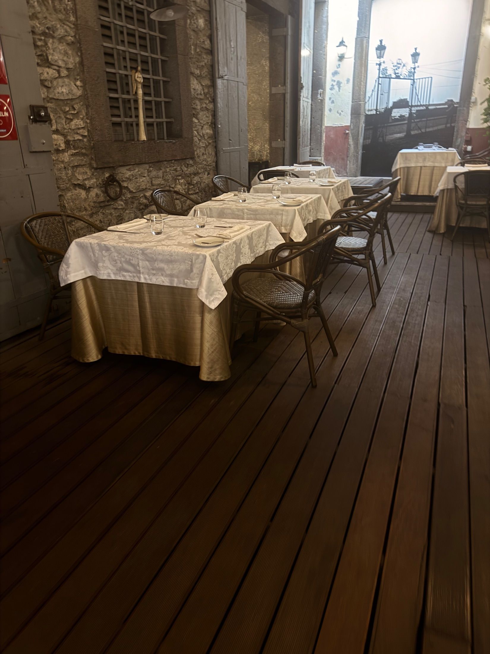Mesas de restaurante com toalhas estampadas e cadeiras de verga junto a uma parede de pedra