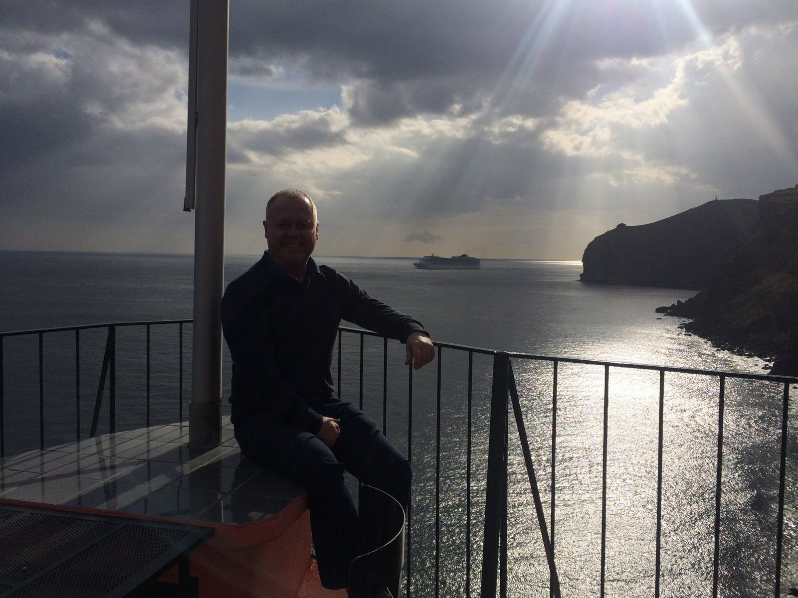 Mike McCabe of Madeira Info at Inn & Art restaurant, Reis Magos, with a cruise ship passing the Atlantic cliffs behind