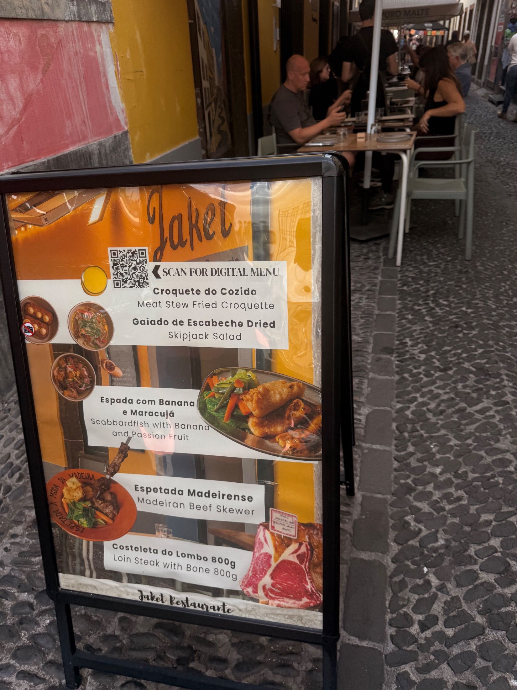 Painel da ementa do restaurante Jaket junto a mesas numa rua de calçada