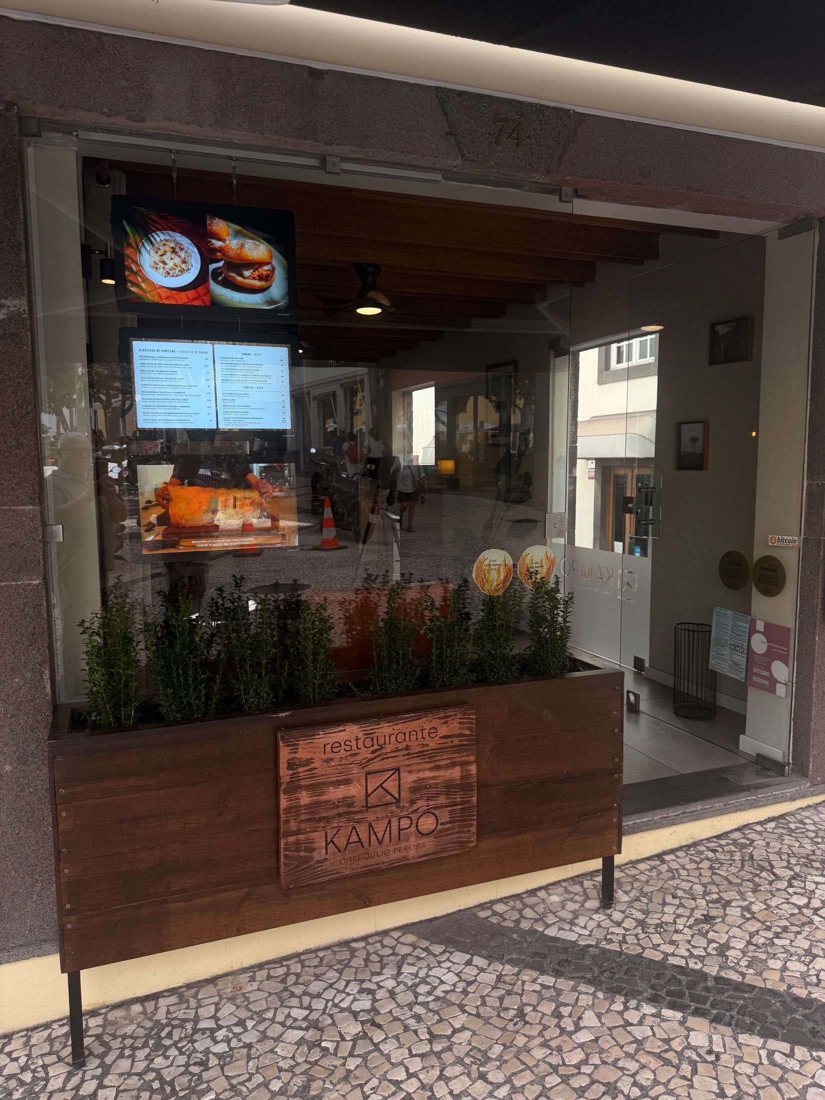 Entrada do restaurante Kampo com portas de vidro, ecrãs de menus e floreira de madeira