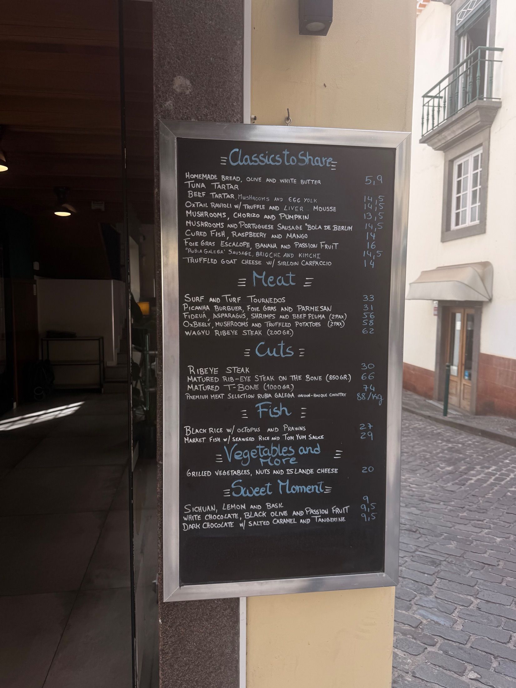 Ementa manuscrita num quadro de ardósia junto à entrada de um restaurante numa rua empedrada