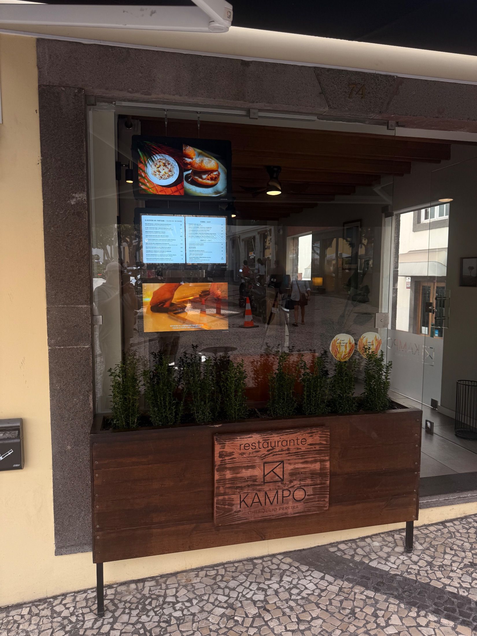 Fachada do restaurante Kampo com floreira de madeira, entrada envidraçada e ecrãs com menu