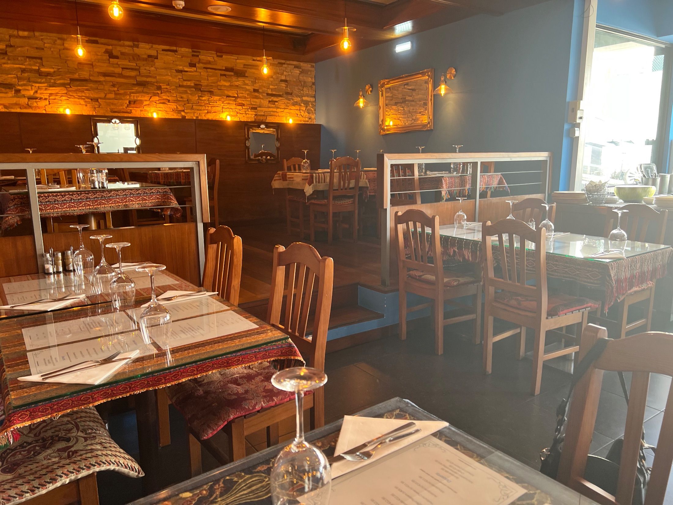 Restaurant interior with patterned tablecloths, wooden chairs, set tables and hanging bulbs
