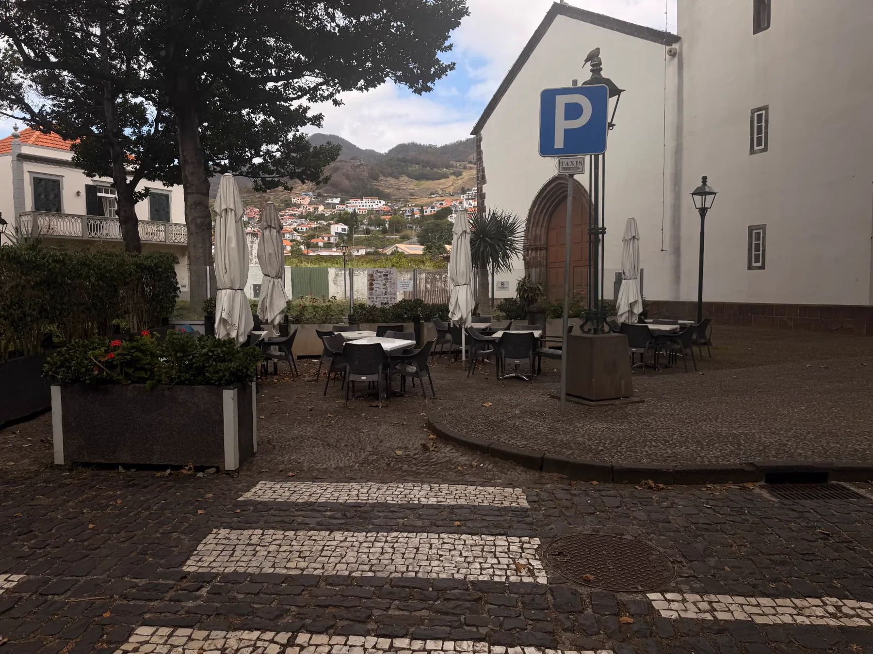 Outdoor café tables beside a white church in Machico, with the hillside homes of the town beyond.
