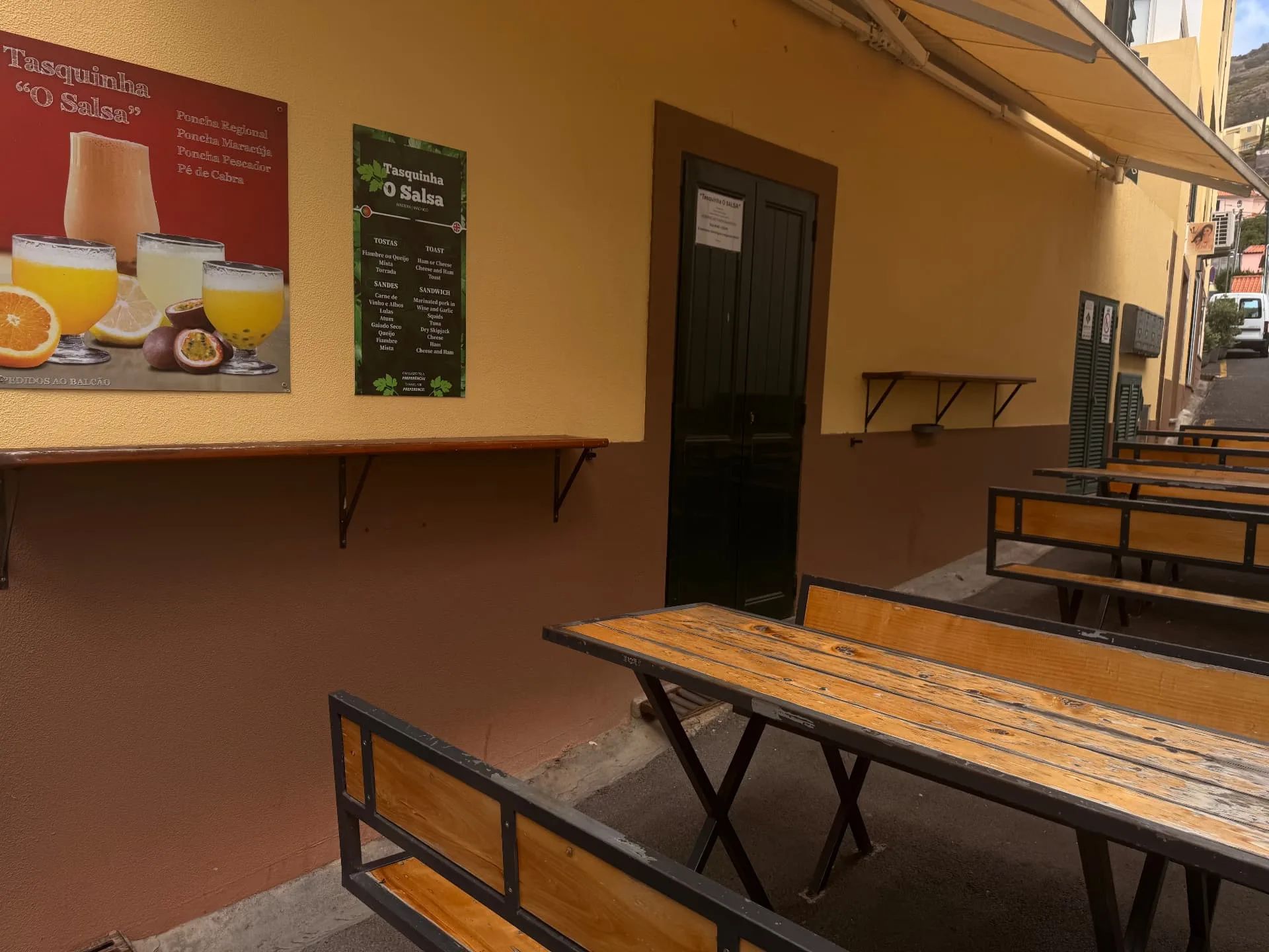 Bancos exteriores na Tasquinha «O Salsa», em Machico, com placas a anunciar poncha e tostas na parede amarela