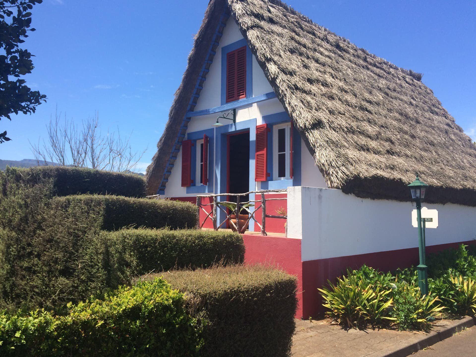 Casa tradicional de Santana, triangular e com telhado de colmo, com persianas vermelhas e acabamentos azuis, com a porta aberta