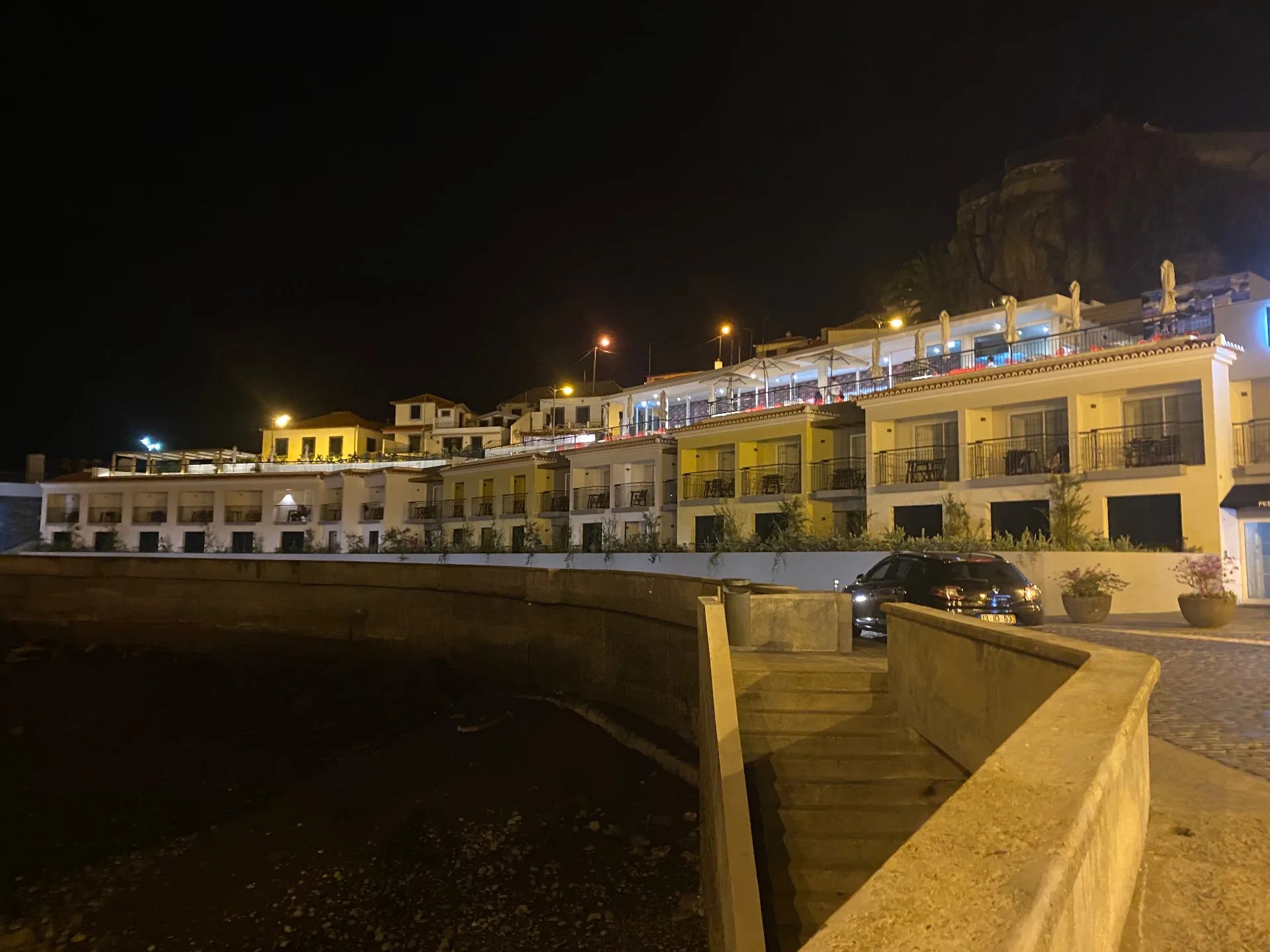 Edifícios da marginal com esplanadas iluminadas à noite, acima da muralha, em Câmara de Lobos